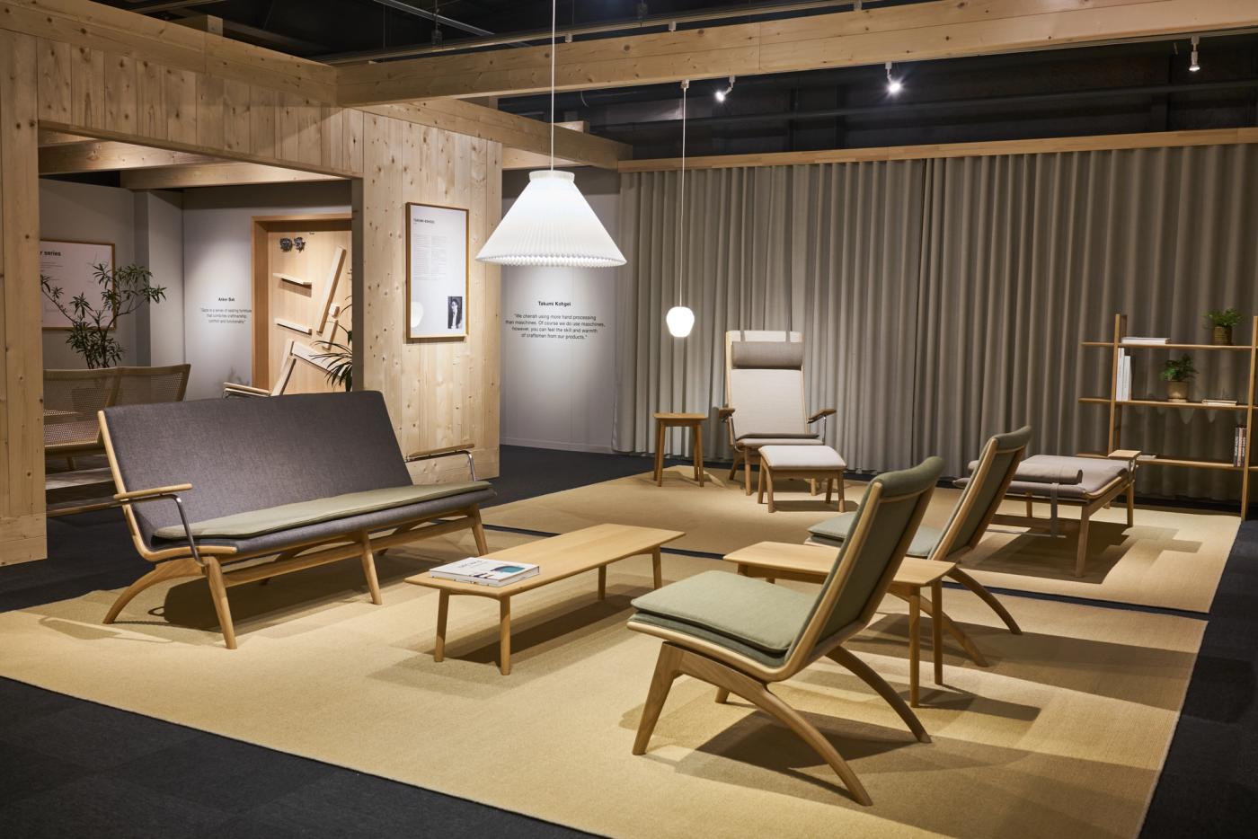 Exhibition room at Gaze in Asahikawa with oak lounge chairs and a sofa with grey and green upholstery, a low oak table, a high-back chair and a daybed on a beige floor, under a white pendant lamp and beside grey curtains.