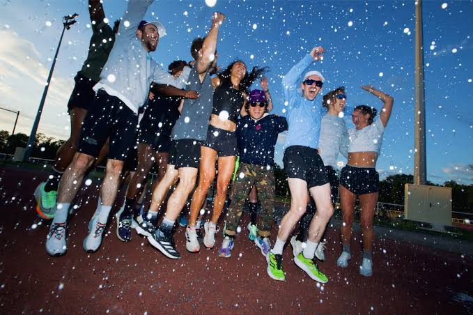 Le crew Jolie Foulée saute de joie sur une piste d’athlétisme, sous une pluie de confettis, au crépuscule.