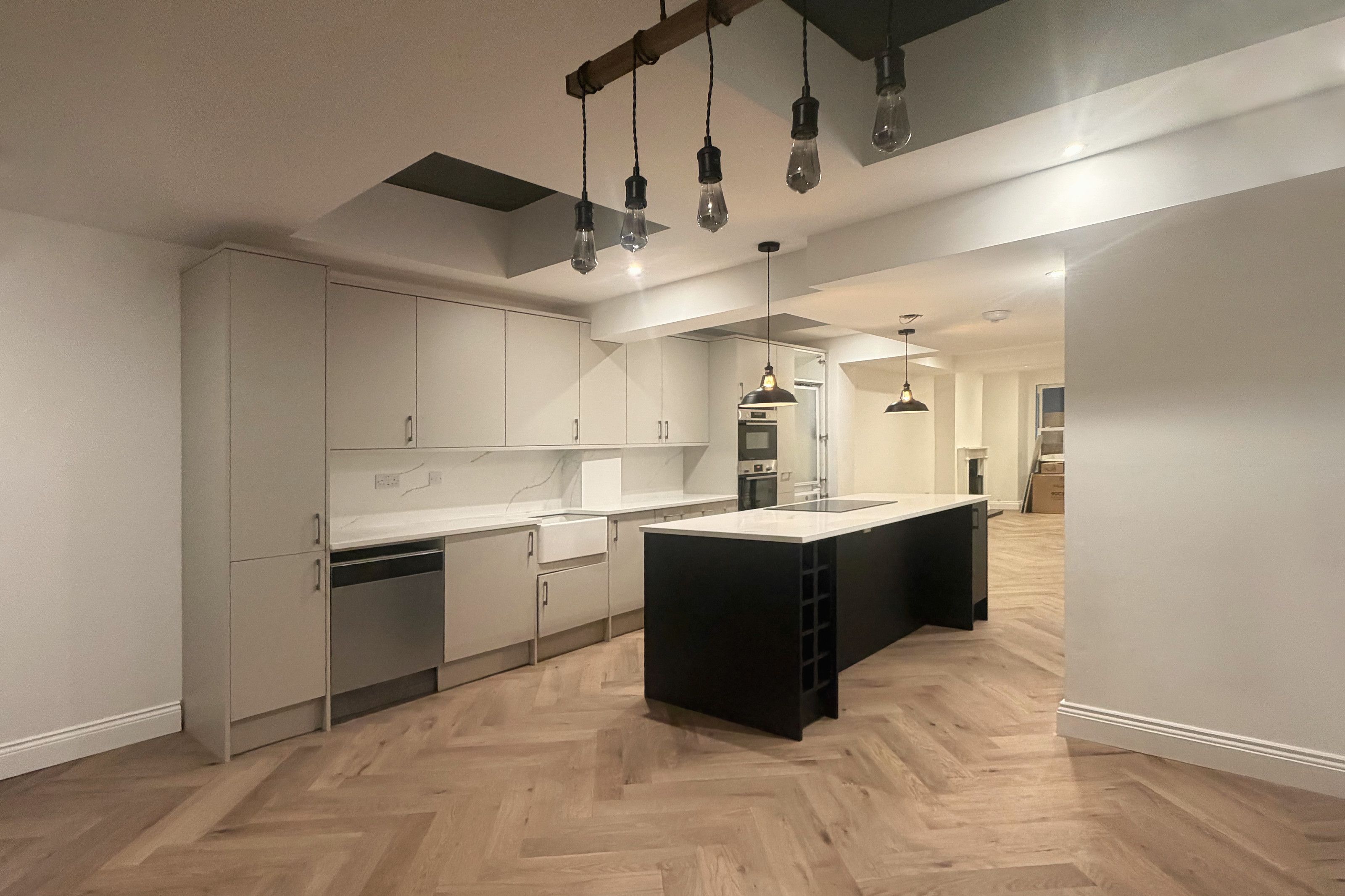 A long kitchen with a black island under pendant lights, grey fitted units and a herringbone wood floor