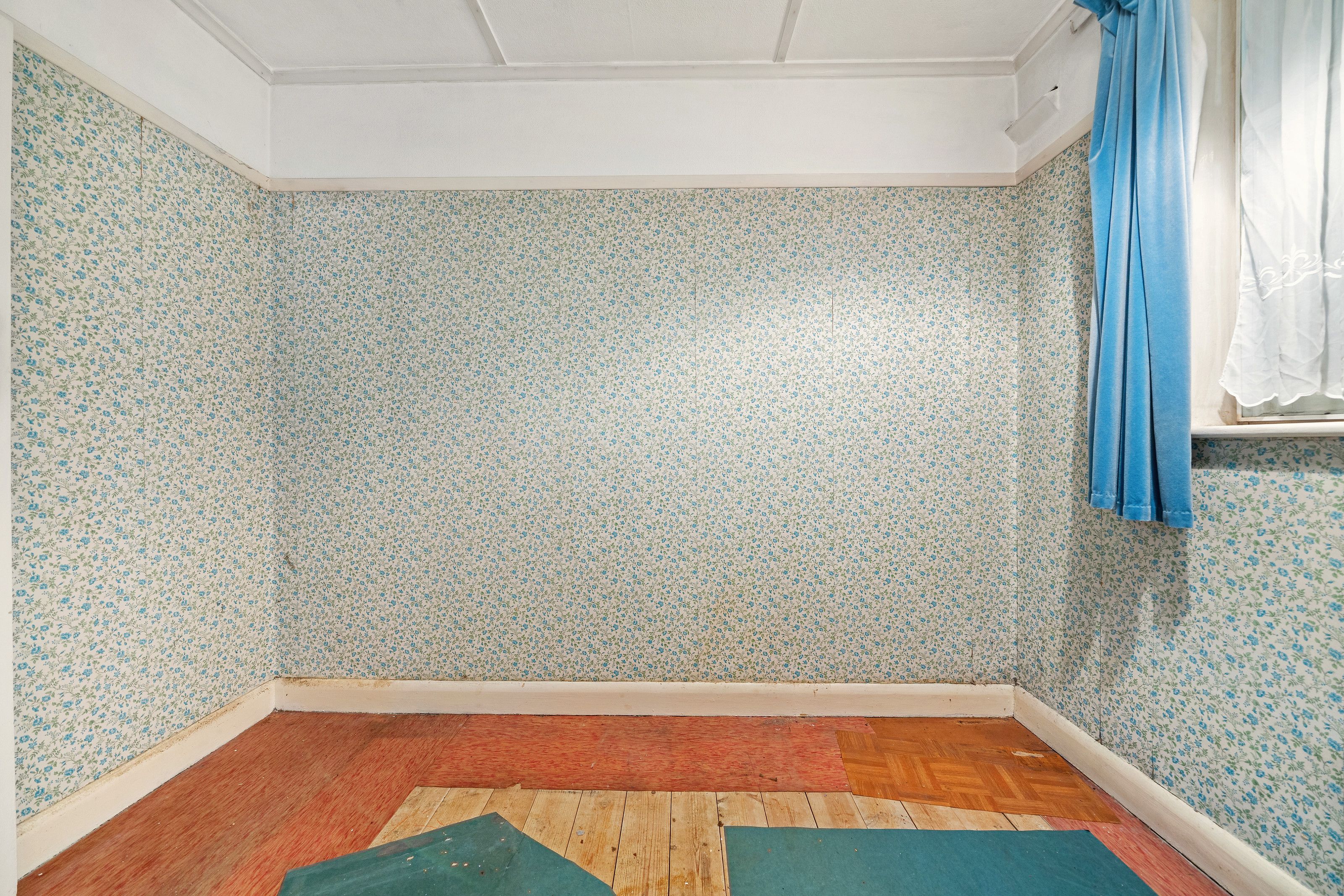 A room before the work, with patterned wallpaper, blue curtains and worn floor covering