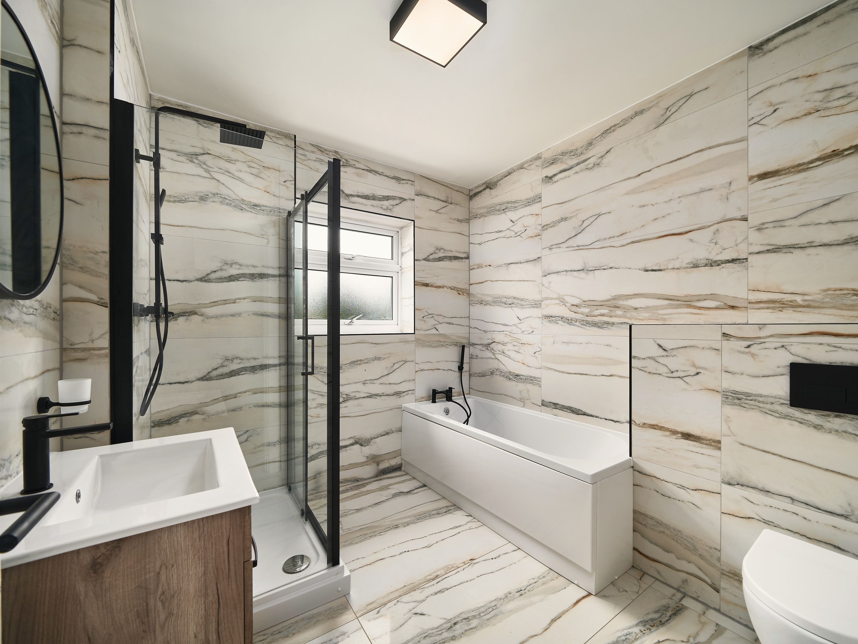 Bathroom tiled floor to ceiling in marble-effect tiles, with a bath, a black-framed shower enclosure and a wood-effect vanity unit