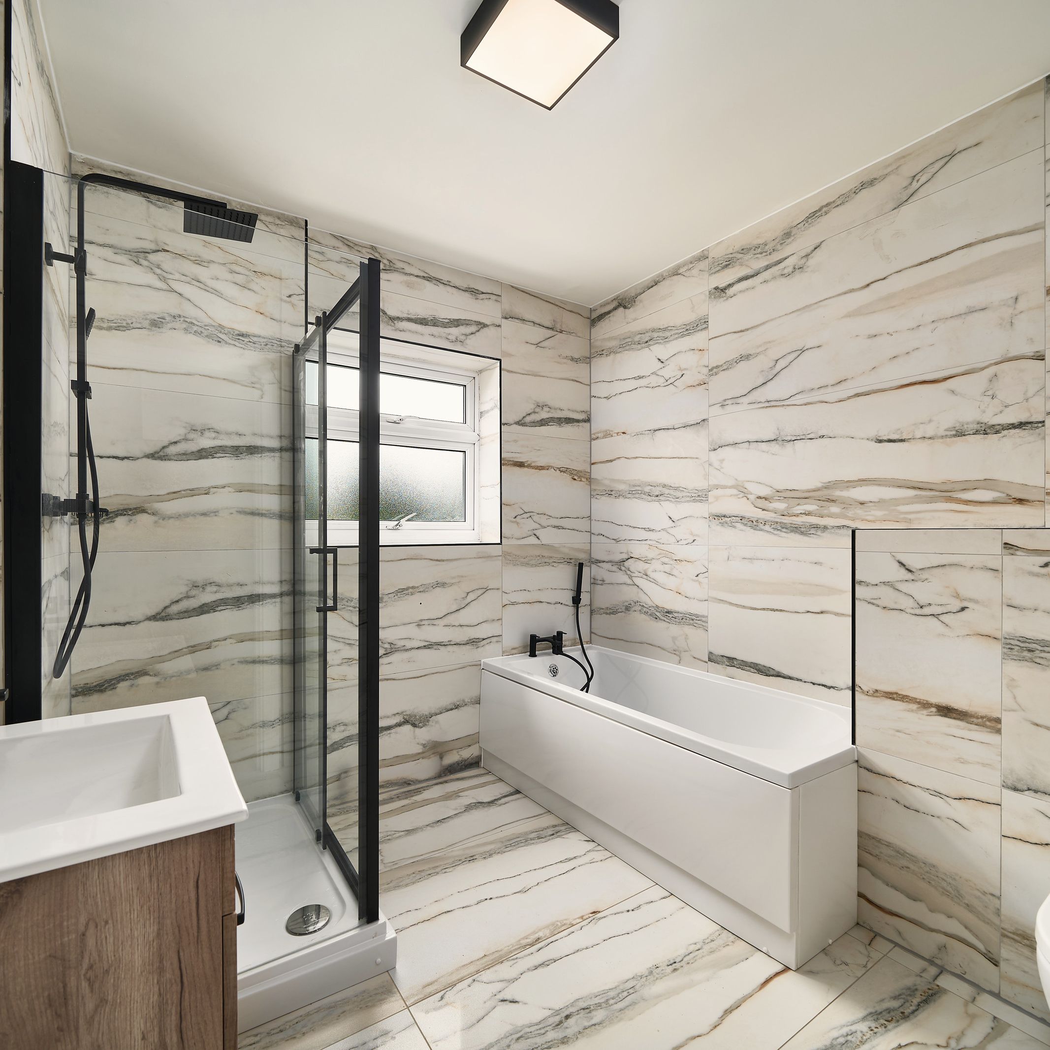 Bathroom tiled floor to ceiling in marble-effect tiles, with a bath, a black-framed shower enclosure and a wood-effect vanity unit