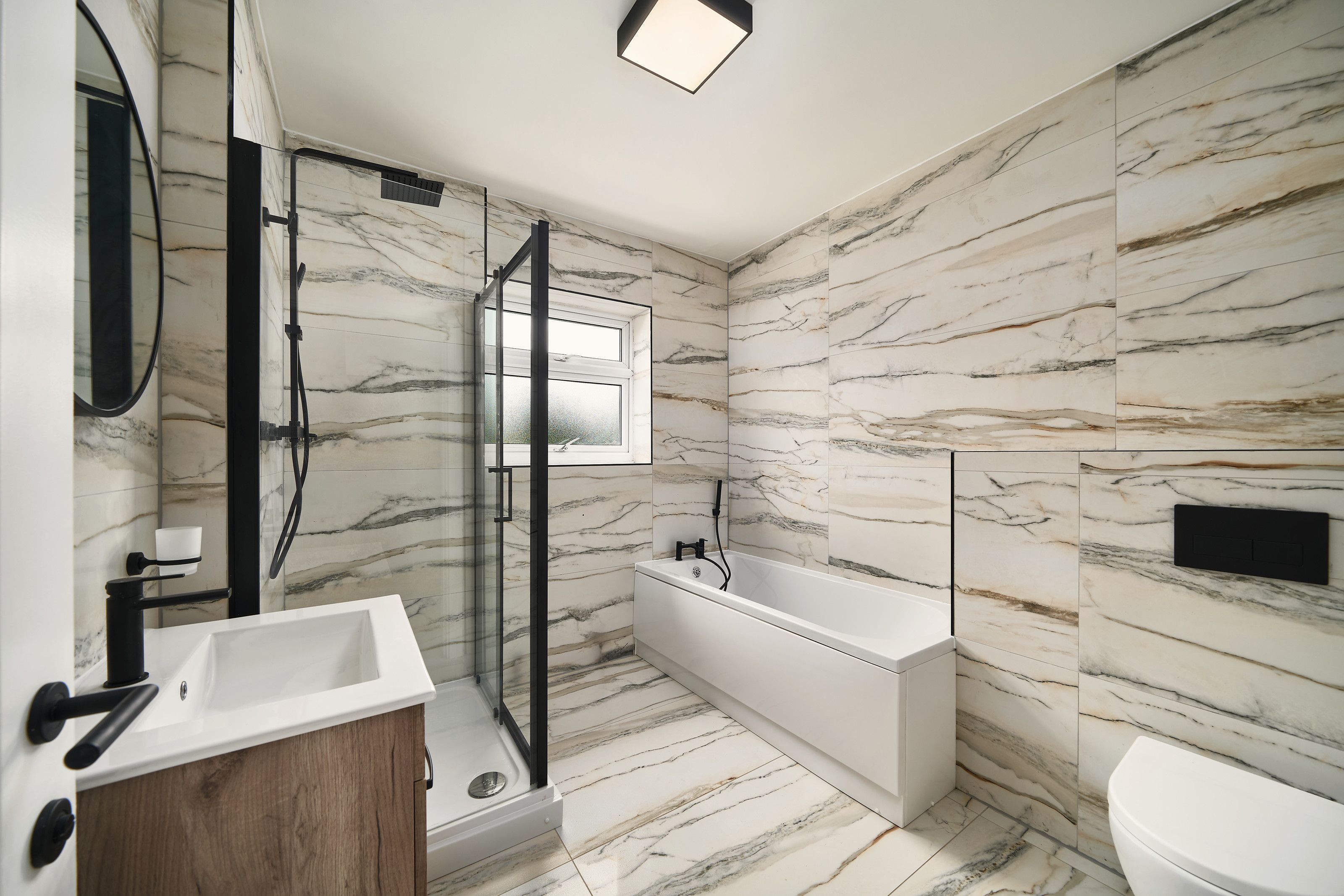 Bathroom tiled floor to ceiling in marble-effect tiles, with a bath, a black-framed shower enclosure and a wood-effect vanity unit