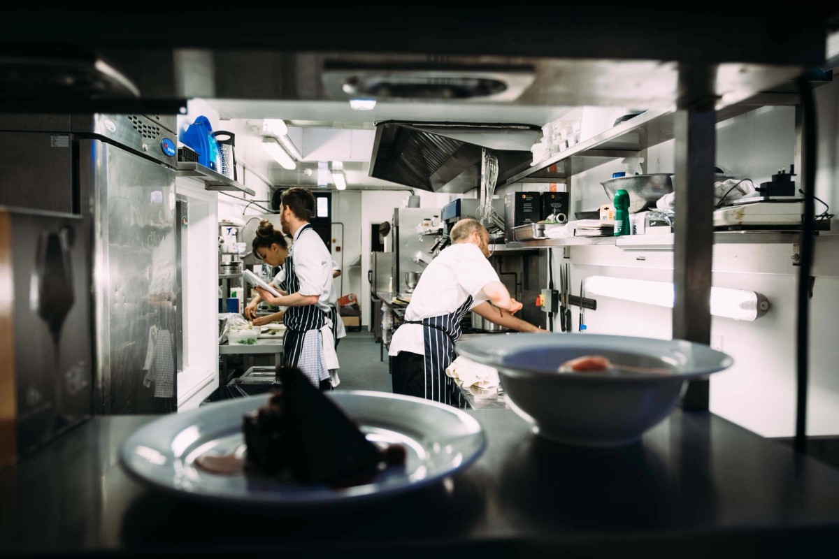 Shot through the kitchen pass with plates waiting in the foreground, the brigade working behind. The controlled intensity of a kitchen in full service.