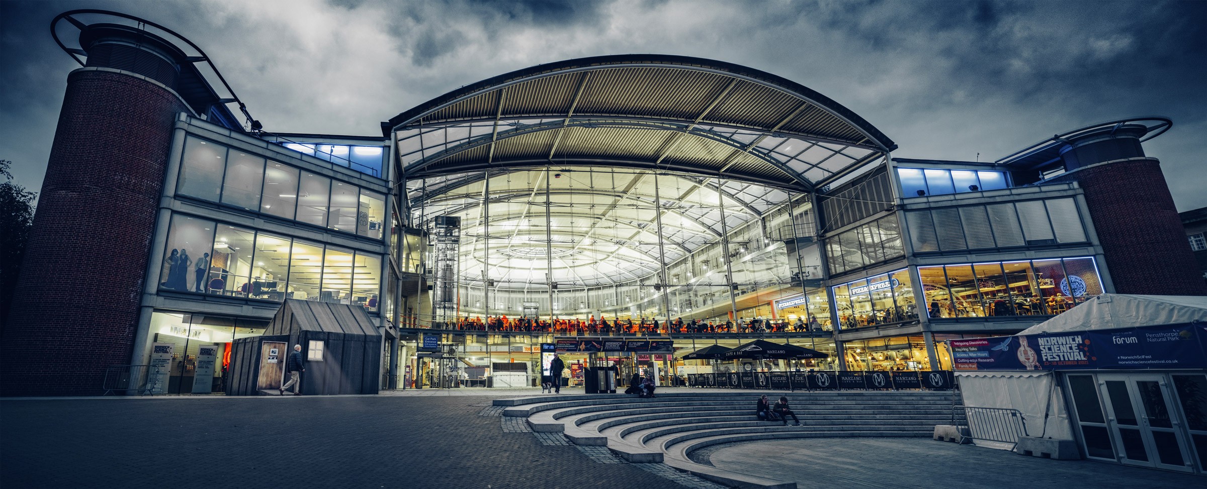 The Forum in Norwich at dusk, its curved glass façade lit from within, framed by brick towers and the amphitheatre steps below.