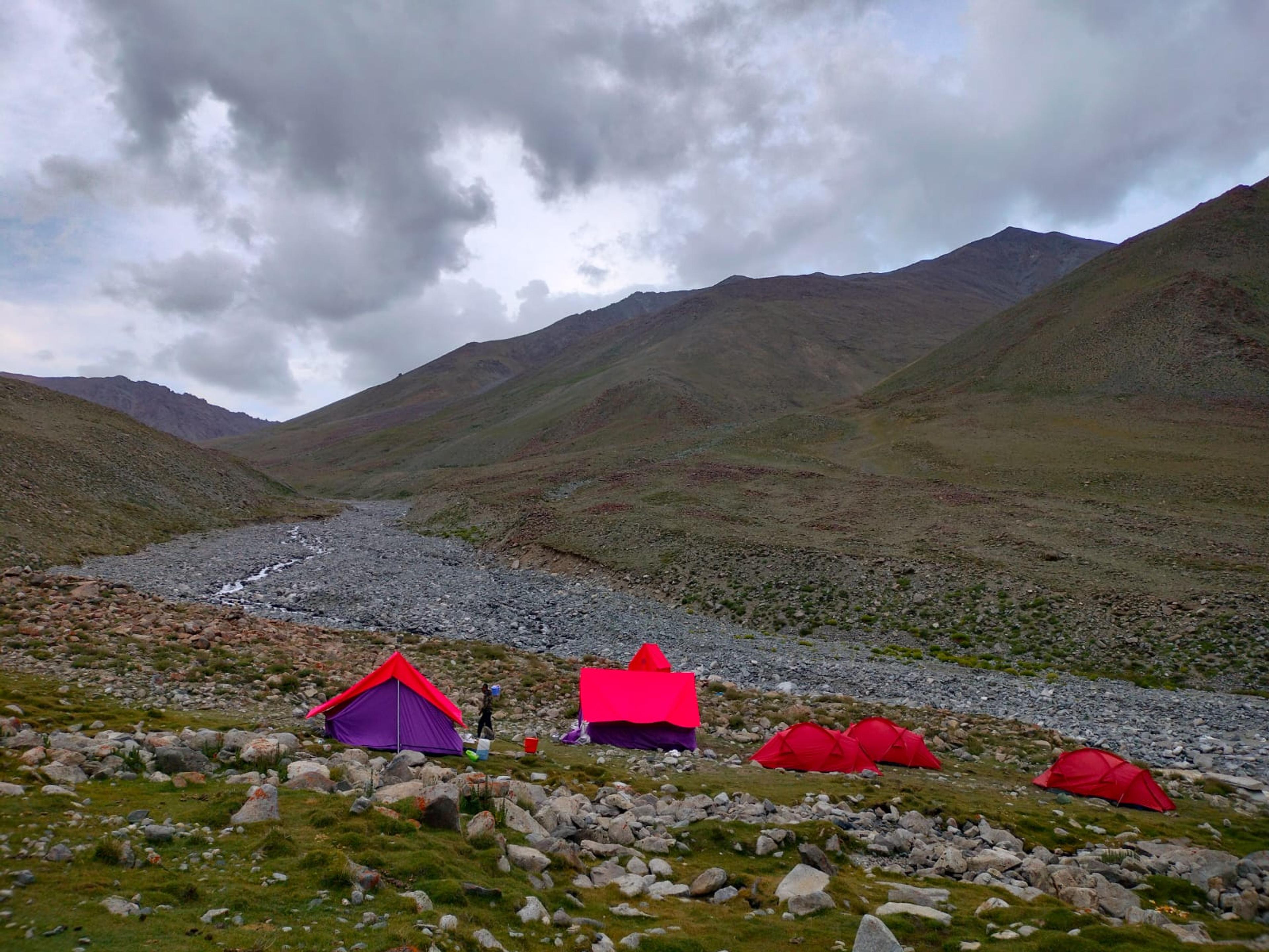 Red sleeping tents and purple-and-pink kitchen tents on a grassy bank beside a rocky stream bed, under heavy clouds