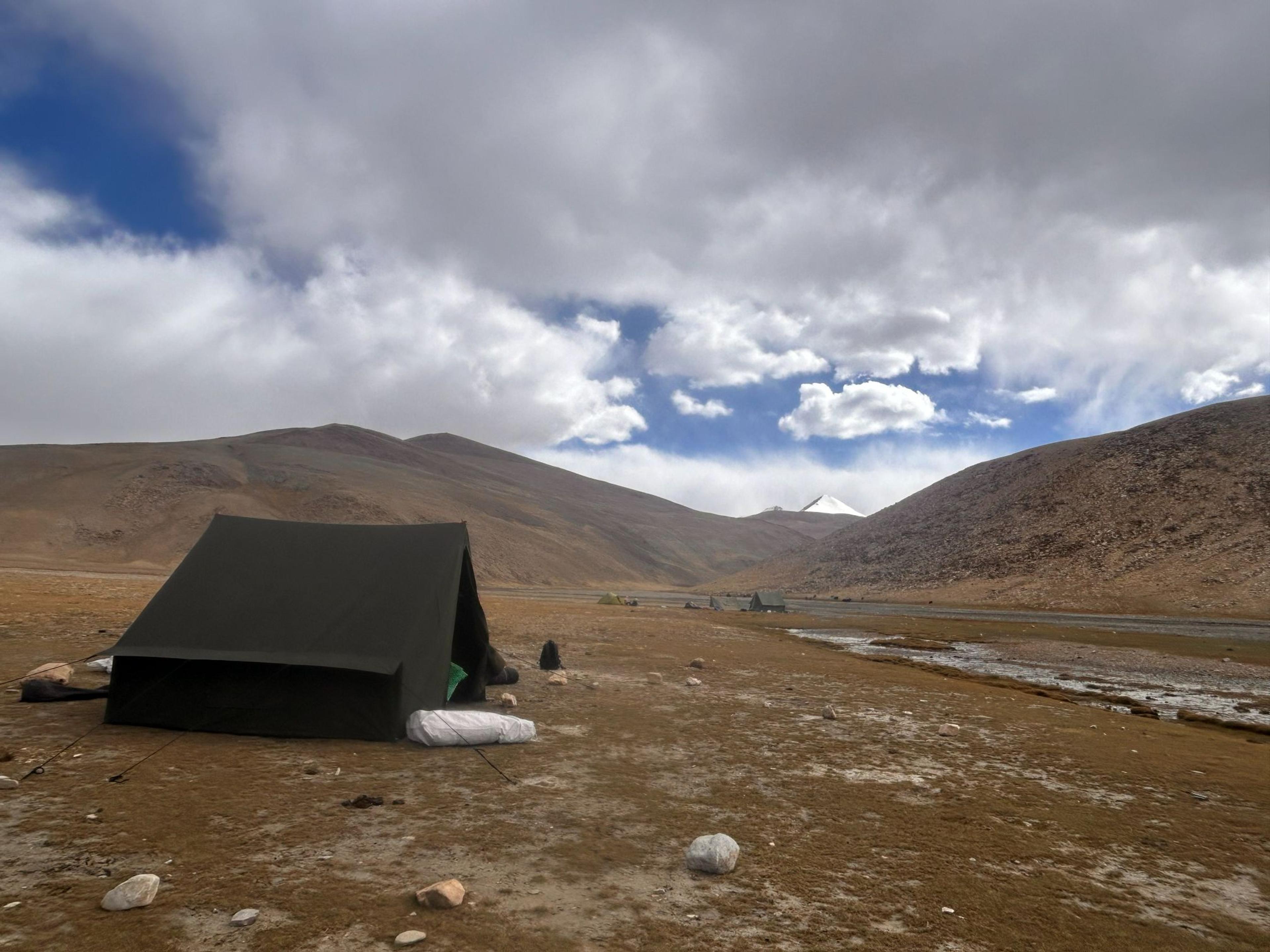 A lone green tent pitched on a barren rocky valley floor near Mentok Kangri base camp with arid brown hills and a glimpse of a snow-capped peak between the mountains in the background, Ladakh