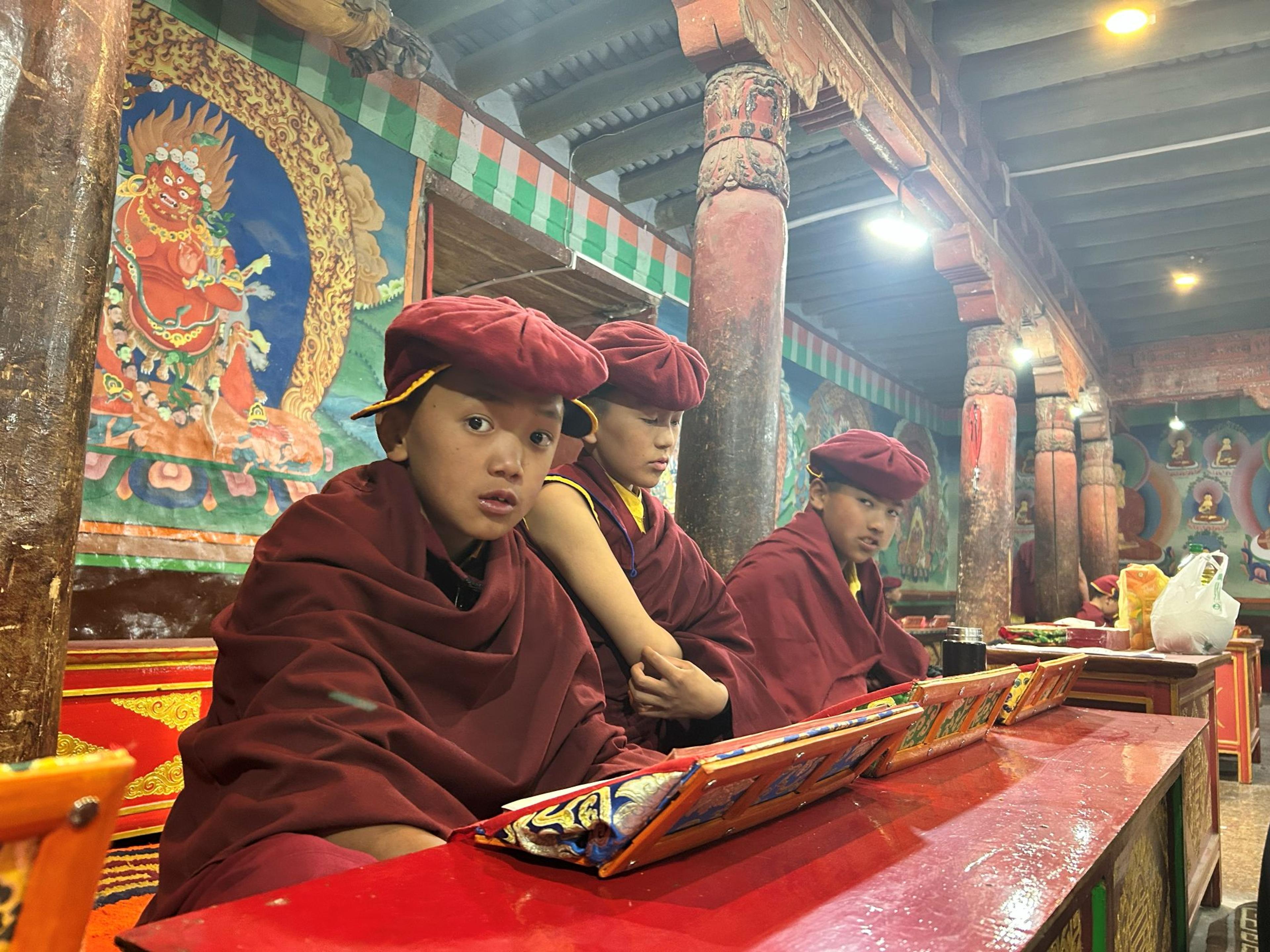 Three young monks in maroon robes and hats seated at a red prayer desk with texts, a painted protector deity on the wall behind