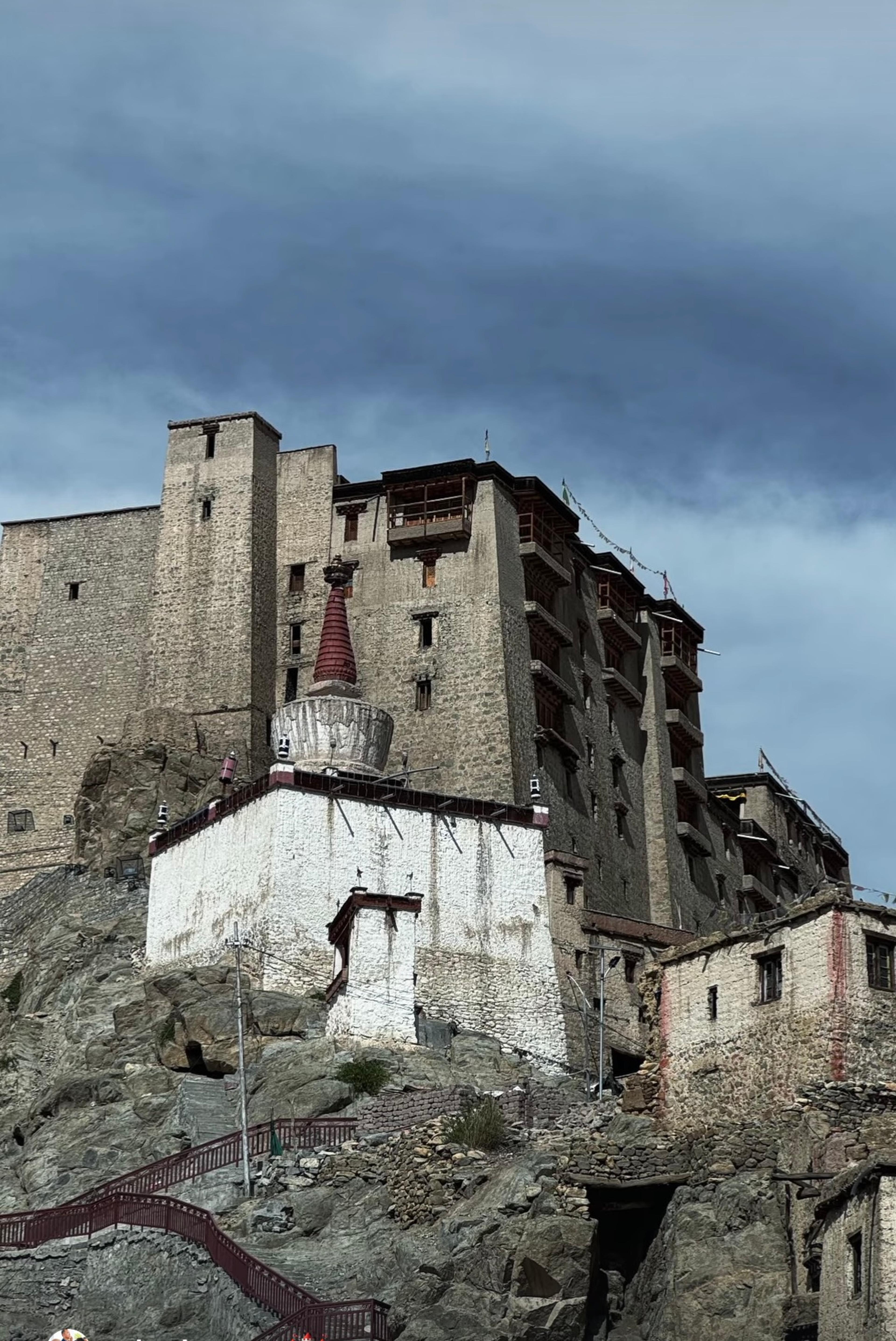 Nine-storey stone Leh Palace rising above a white chorten and rocky hillside, under dark storm clouds