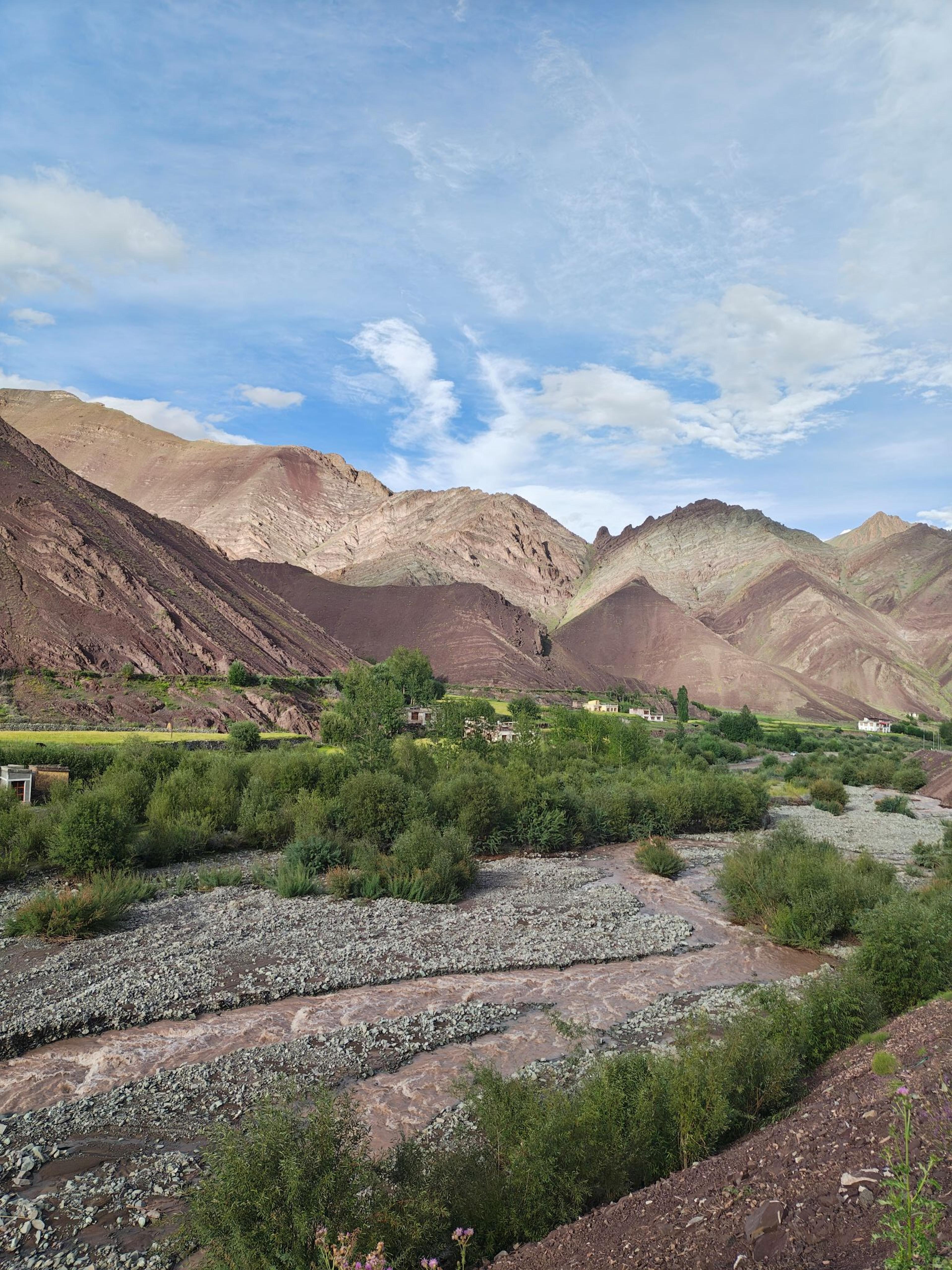 Braided stream through willow scrub and green fields, with village houses below maroon and pink striped mountains