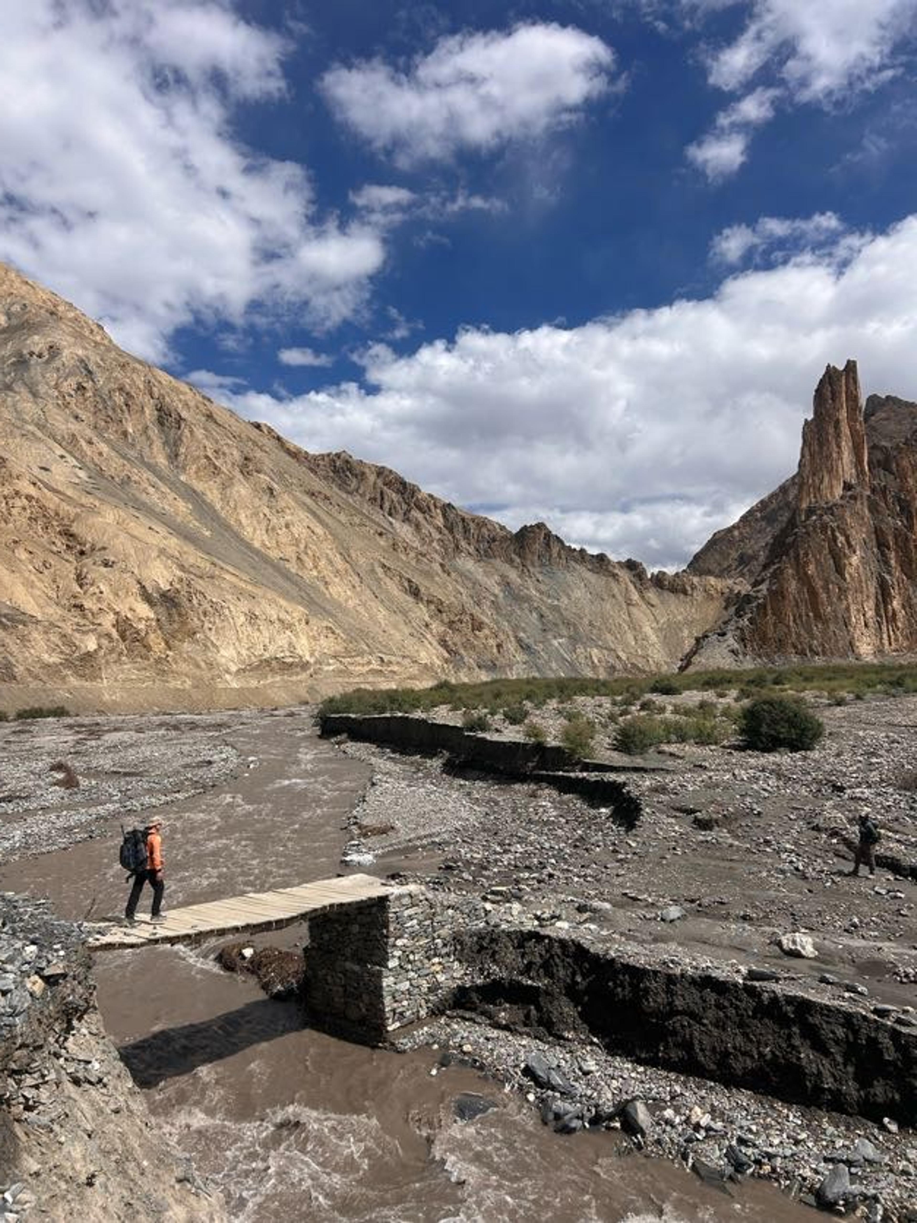 A trekker with an orange backpack crossing a narrow wooden plank bridge over a rushing glacial river in Markha Valley with dramatic sandstone spires and canyon walls in the background, Ladakh
