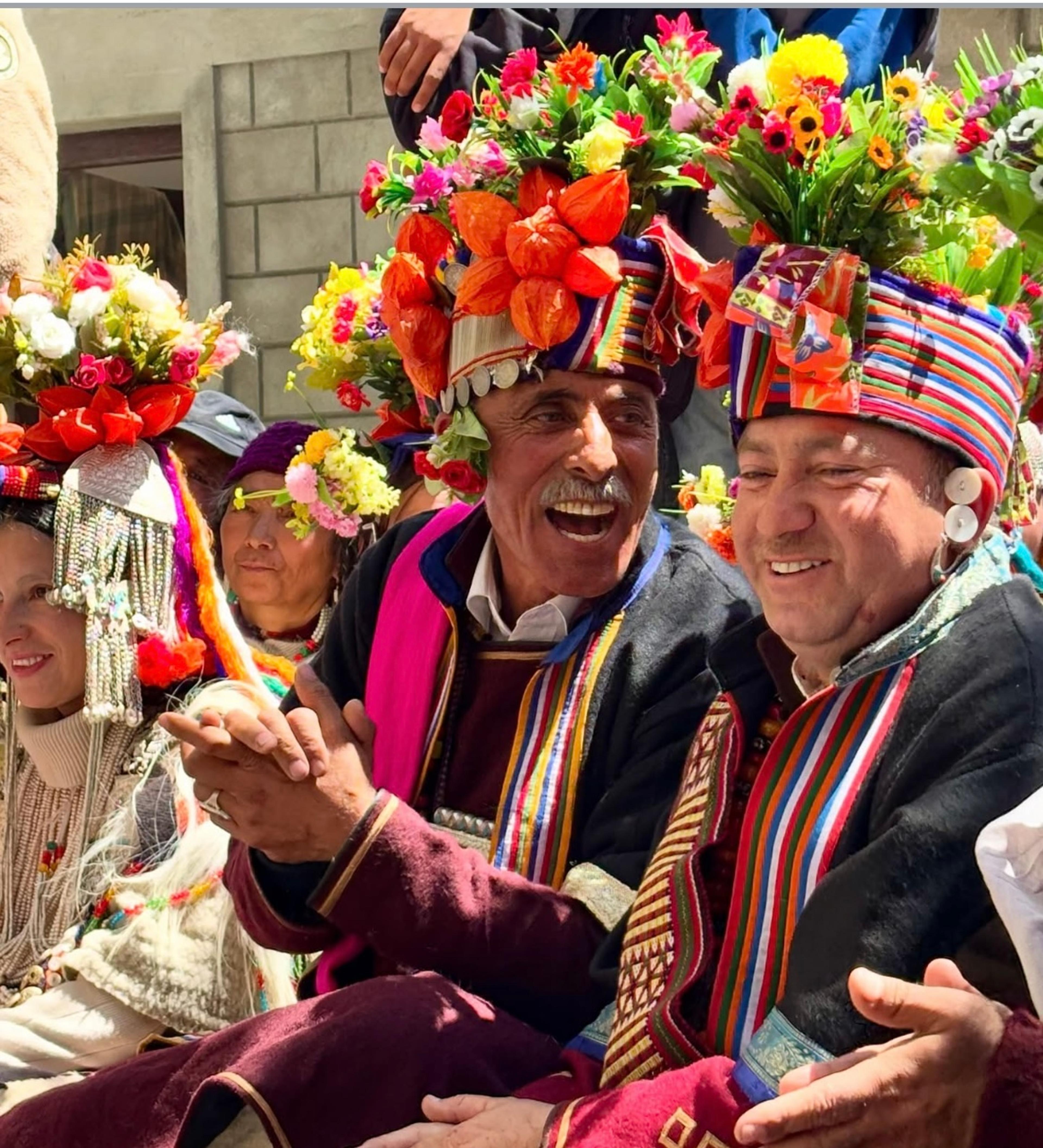 Brokpa men and women in traditional dress and tall flower-covered headdresses, smiling and clapping at a celebration