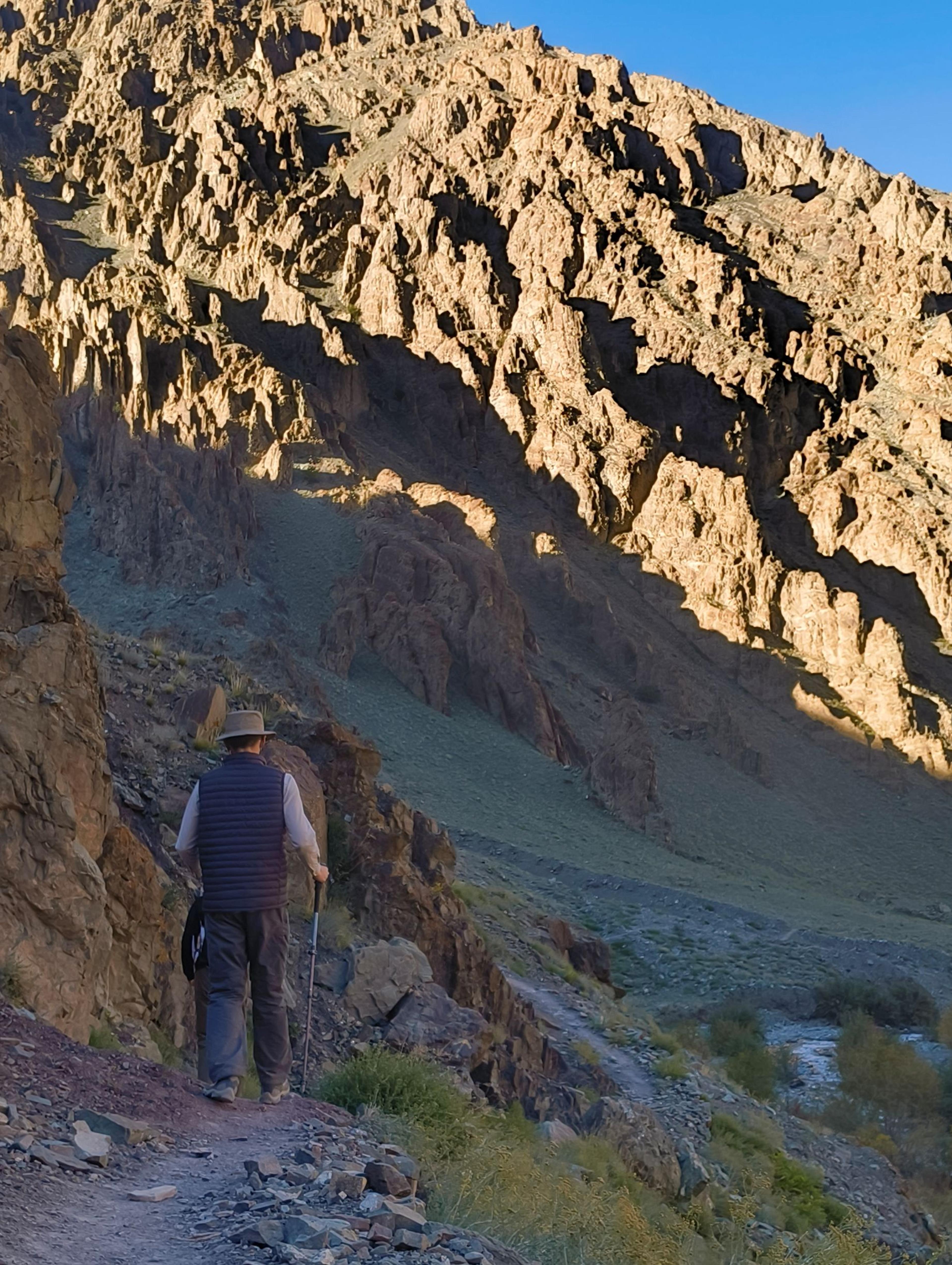 Hiker with a walking pole on a rocky trail in a gorge, sunlit jagged cliffs above and a stream below