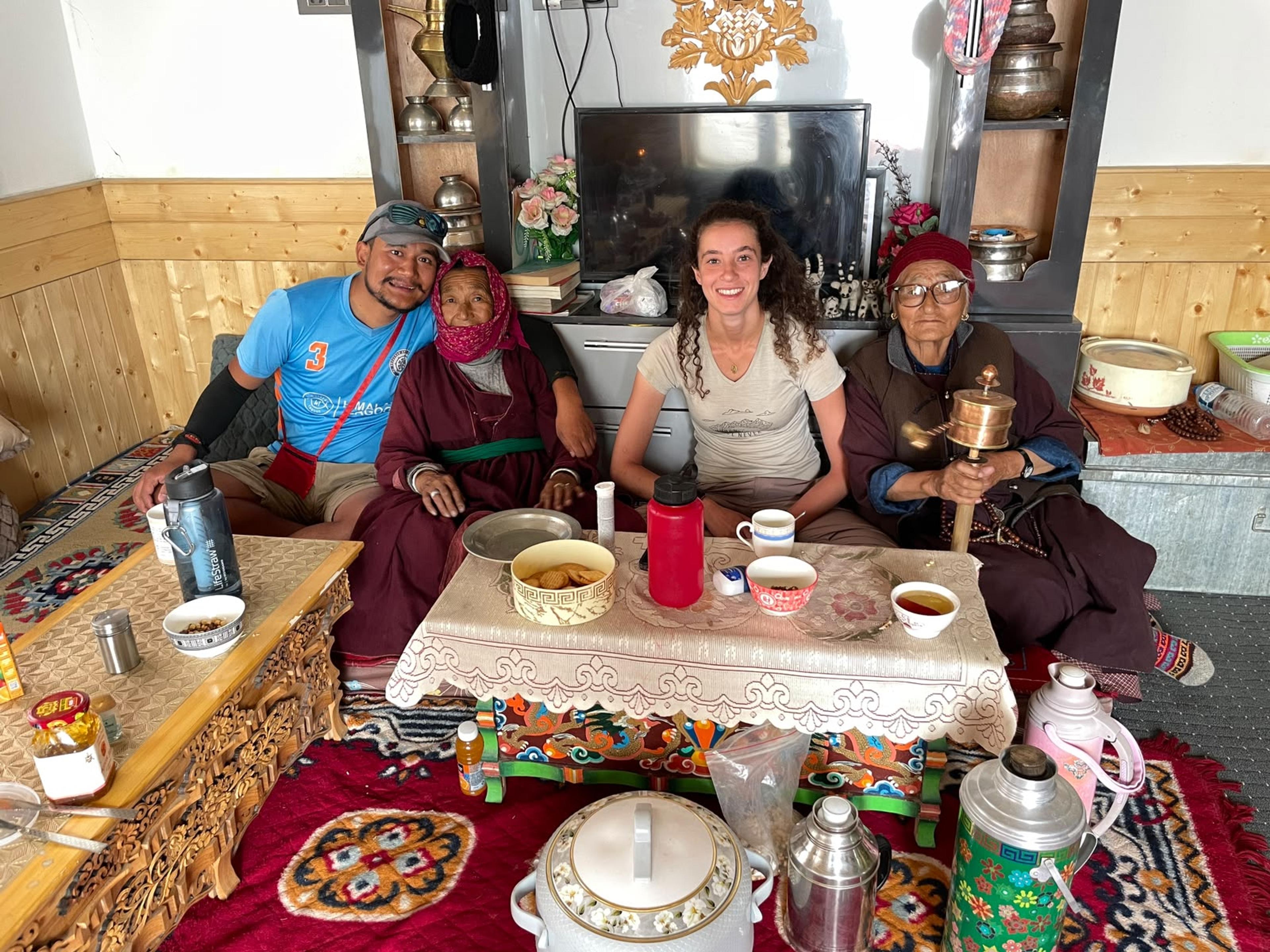 Two trekkers having tea with two elderly Ladakhi women in traditional dress, one holding a prayer wheel, in a homestay