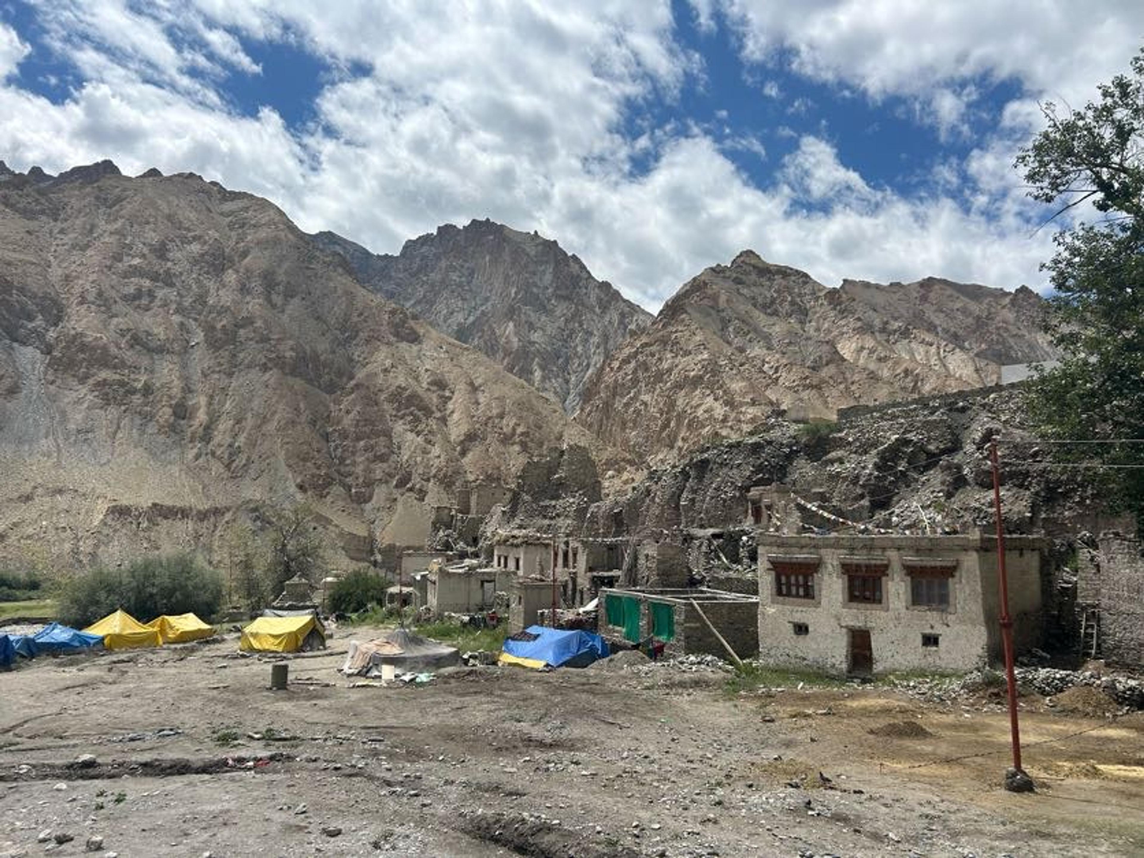 Traditional stone houses and colorful trekking tents set up alongside a remote Ladakhi village in Markha Valley with towering rugged mountain cliffs rising directly behind