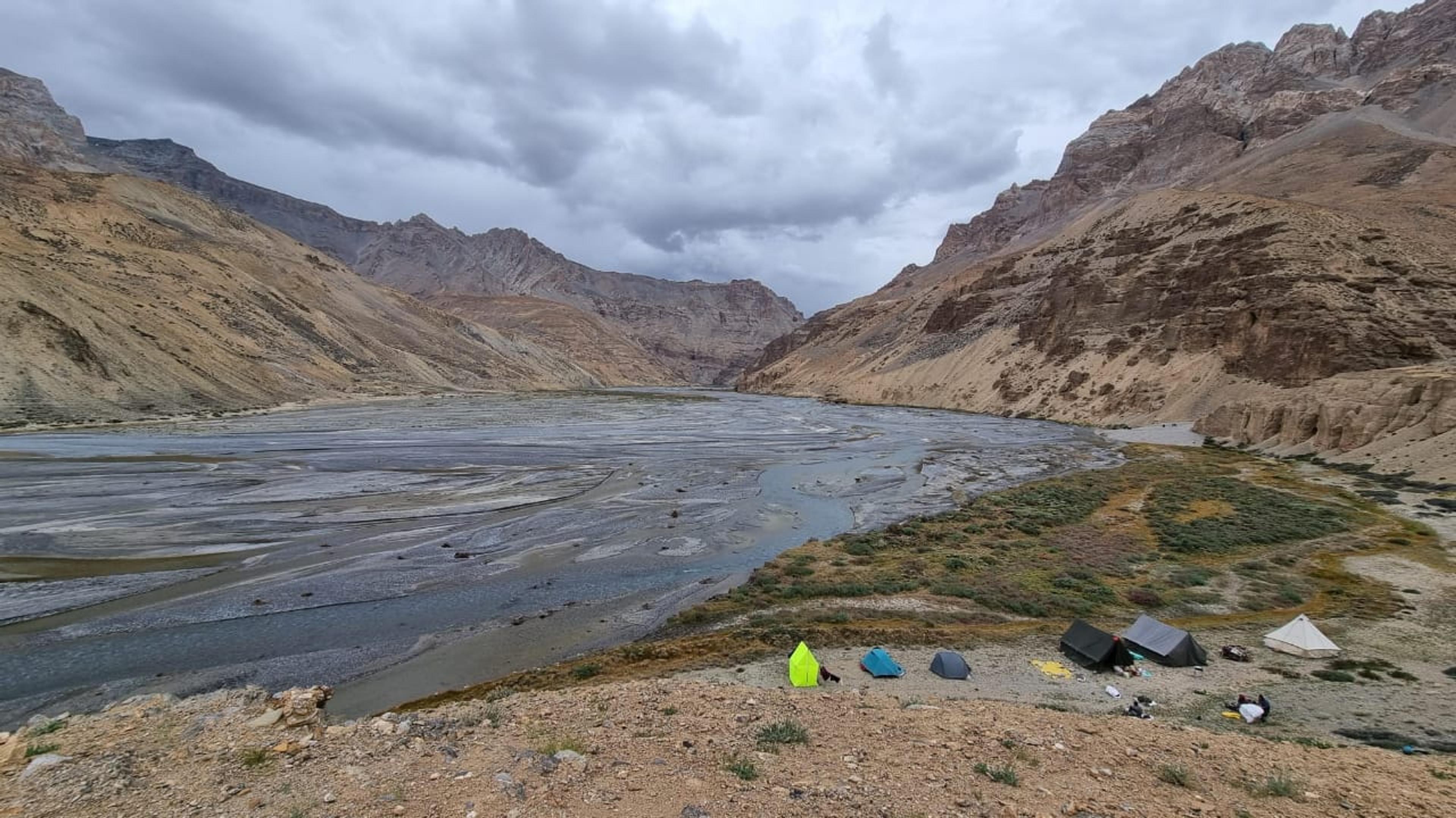 Small camp of tents on a grassy bank beside a wide braided river in a deep, arid valley under grey clouds