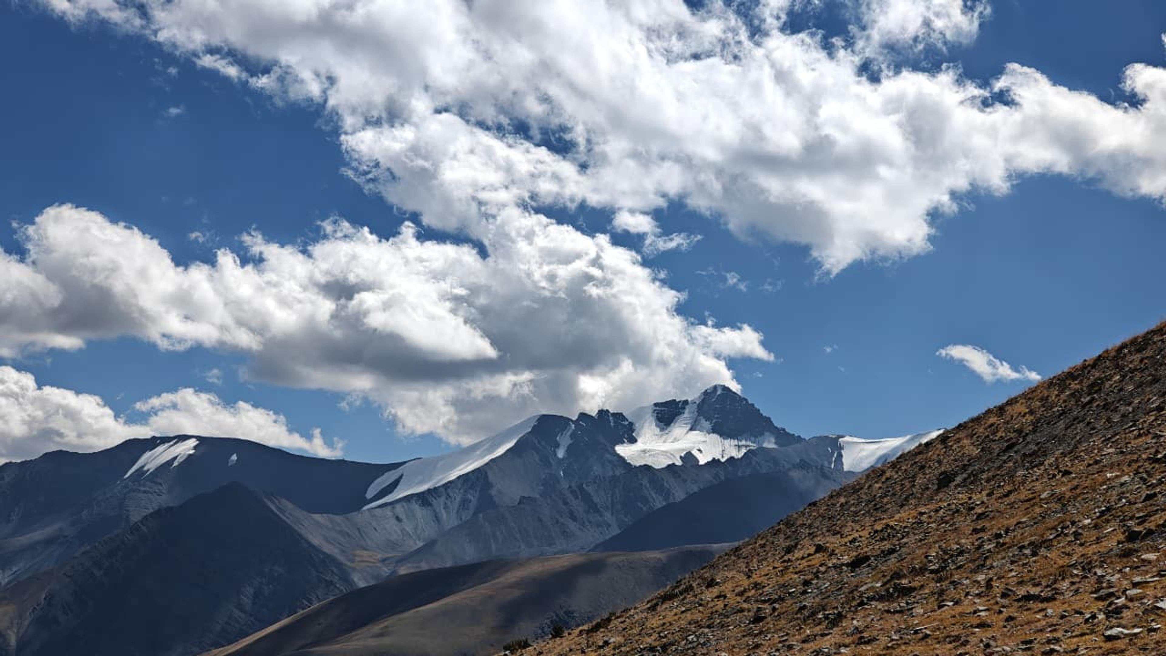 Snow-capped peak with small glaciers above dark ridges, a dry grassy slope in front and white clouds in a blue sky