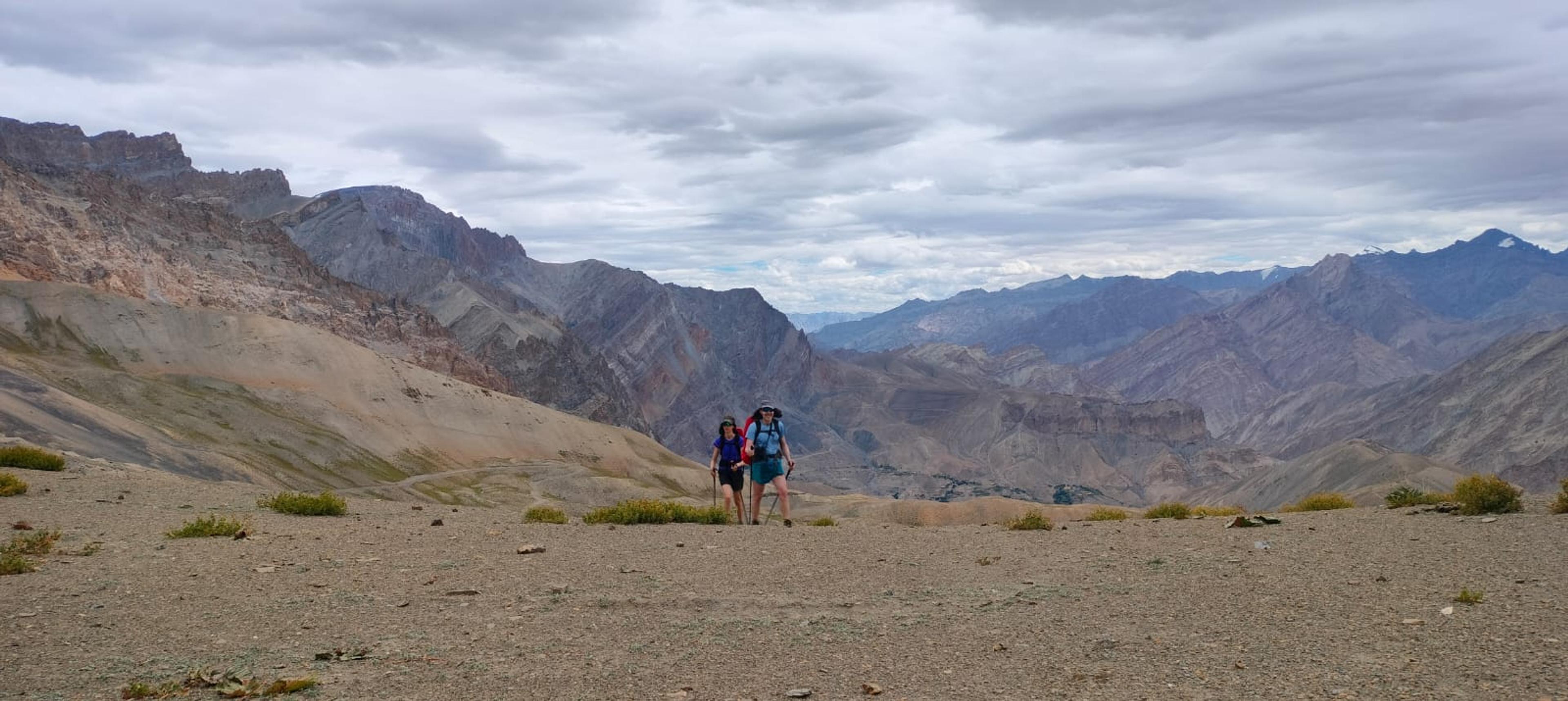 Two trekkers with poles and backpacks on a high gravel shoulder, with layered, jagged mountain ranges behind under clouds