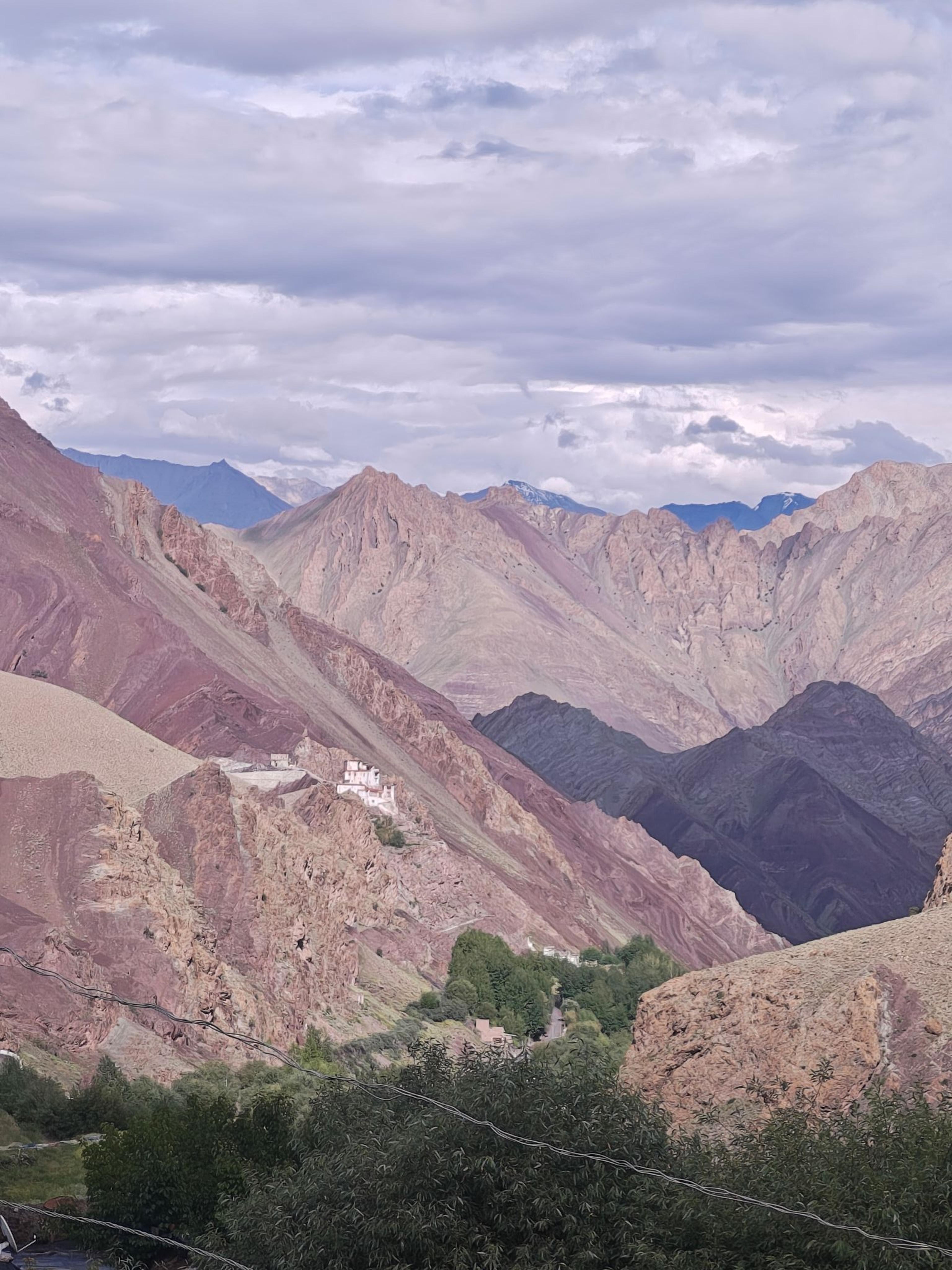 Small white monastery perched on a red rock ridge above a green village oasis, layered pink and purple mountains behind