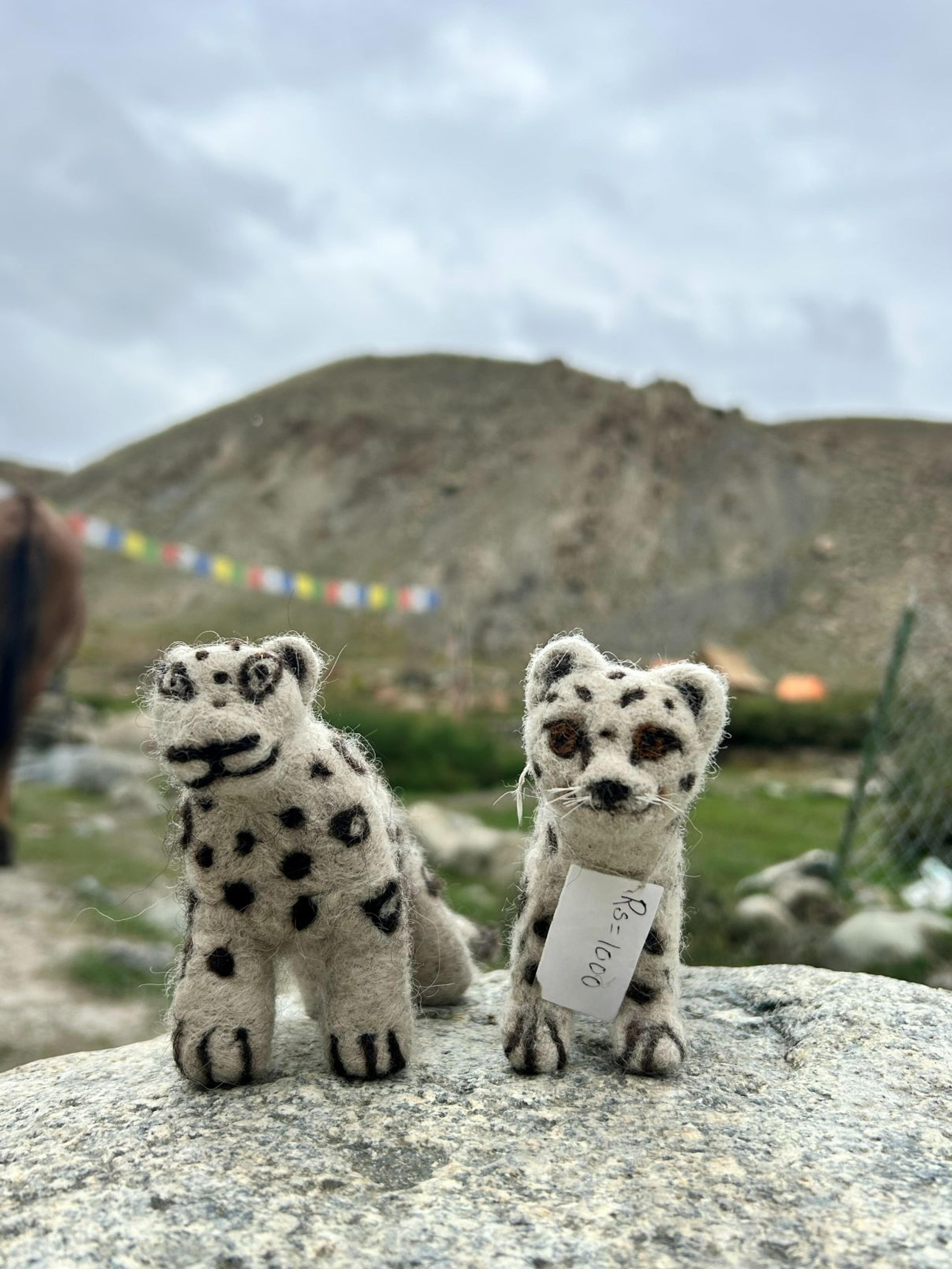 Two handmade felted wool snow leopard toys on a rock, prayer flags and a hillside in the background