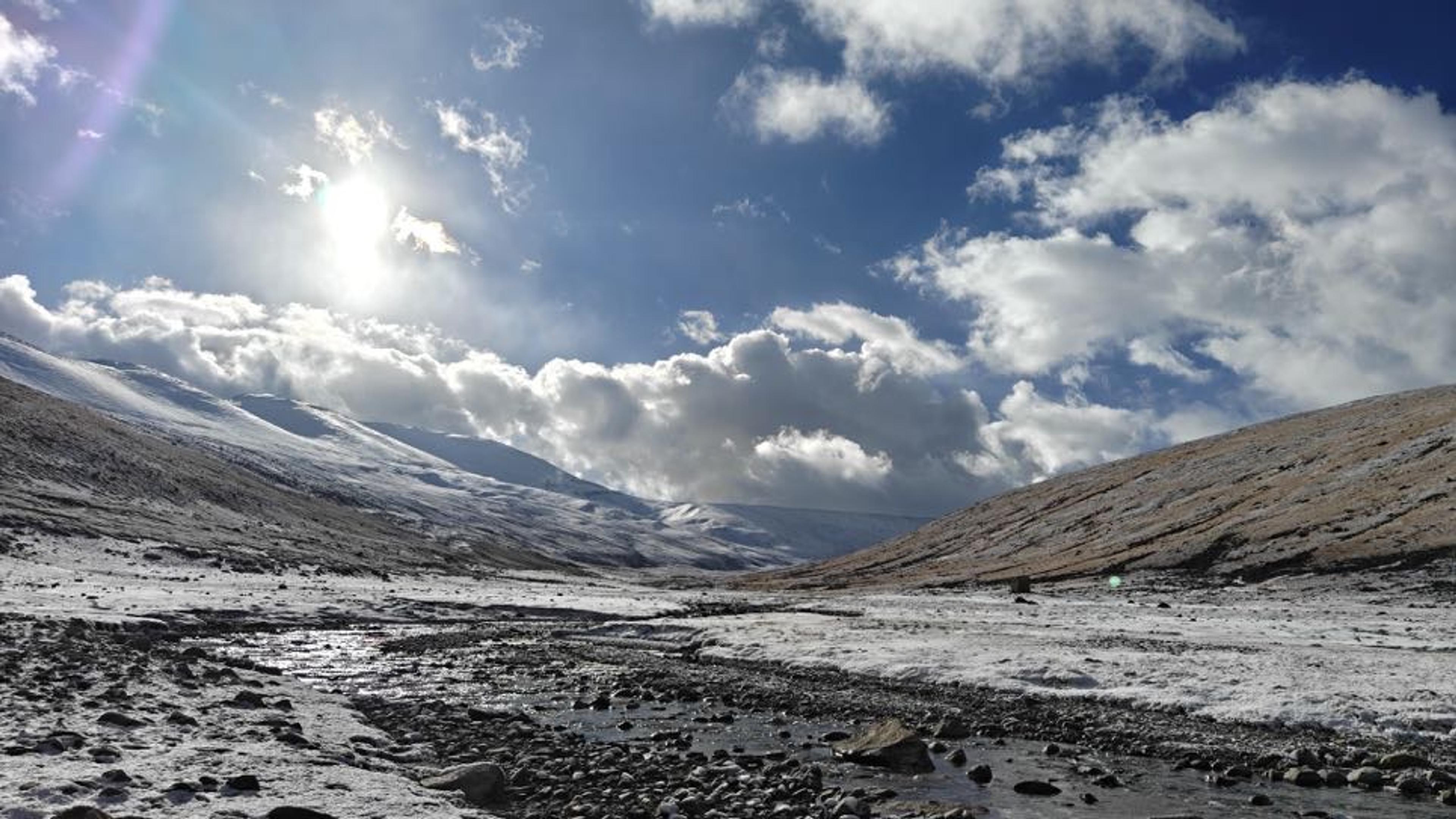 Sun-lit frozen stream running through a snow-covered high-altitude valley in Ladakh with rugged hillsides and dramatic clouds