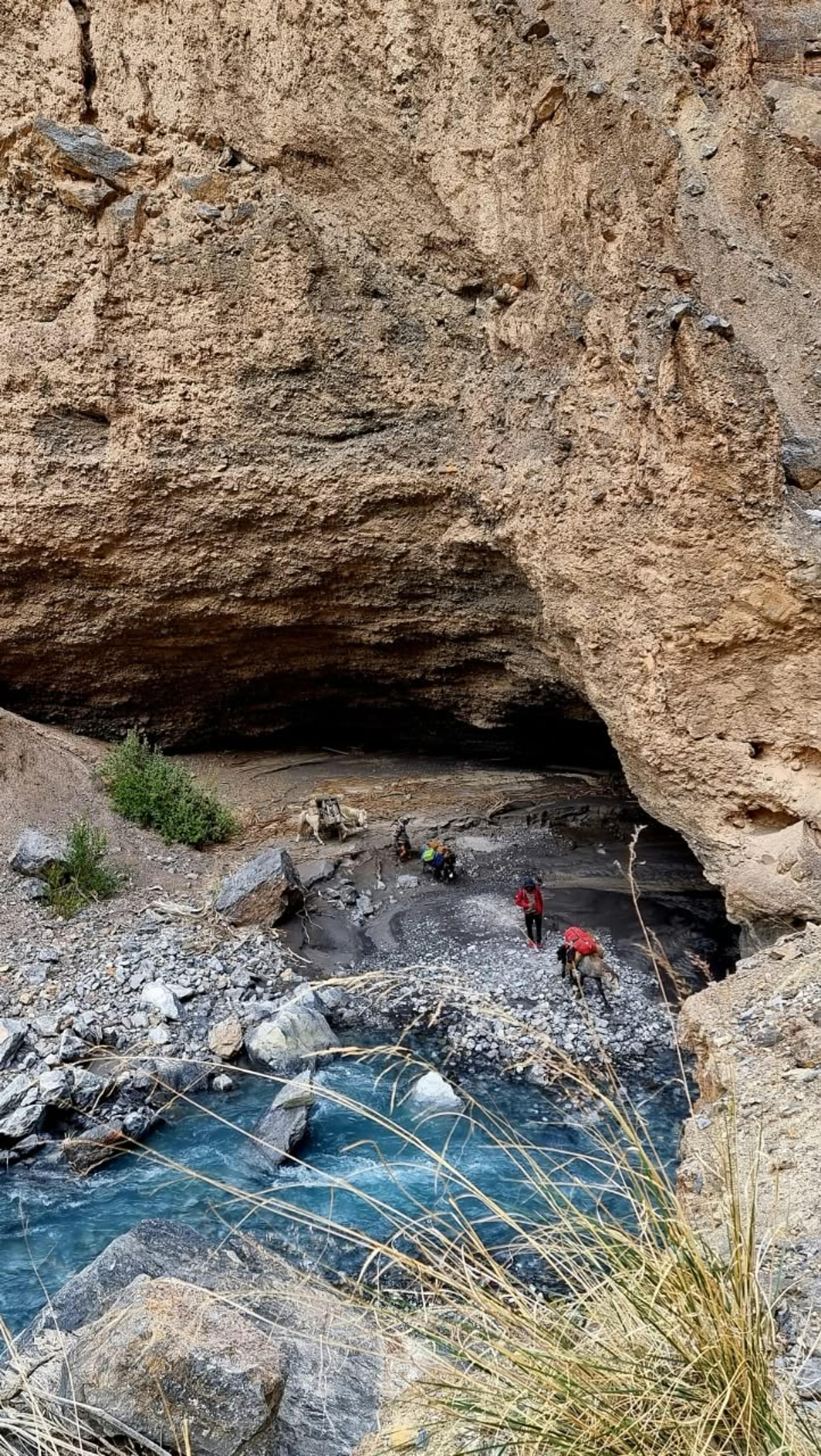 Trekkers and loaded pack horses passing beneath a huge rock overhang beside a turquoise stream in a narrow canyon