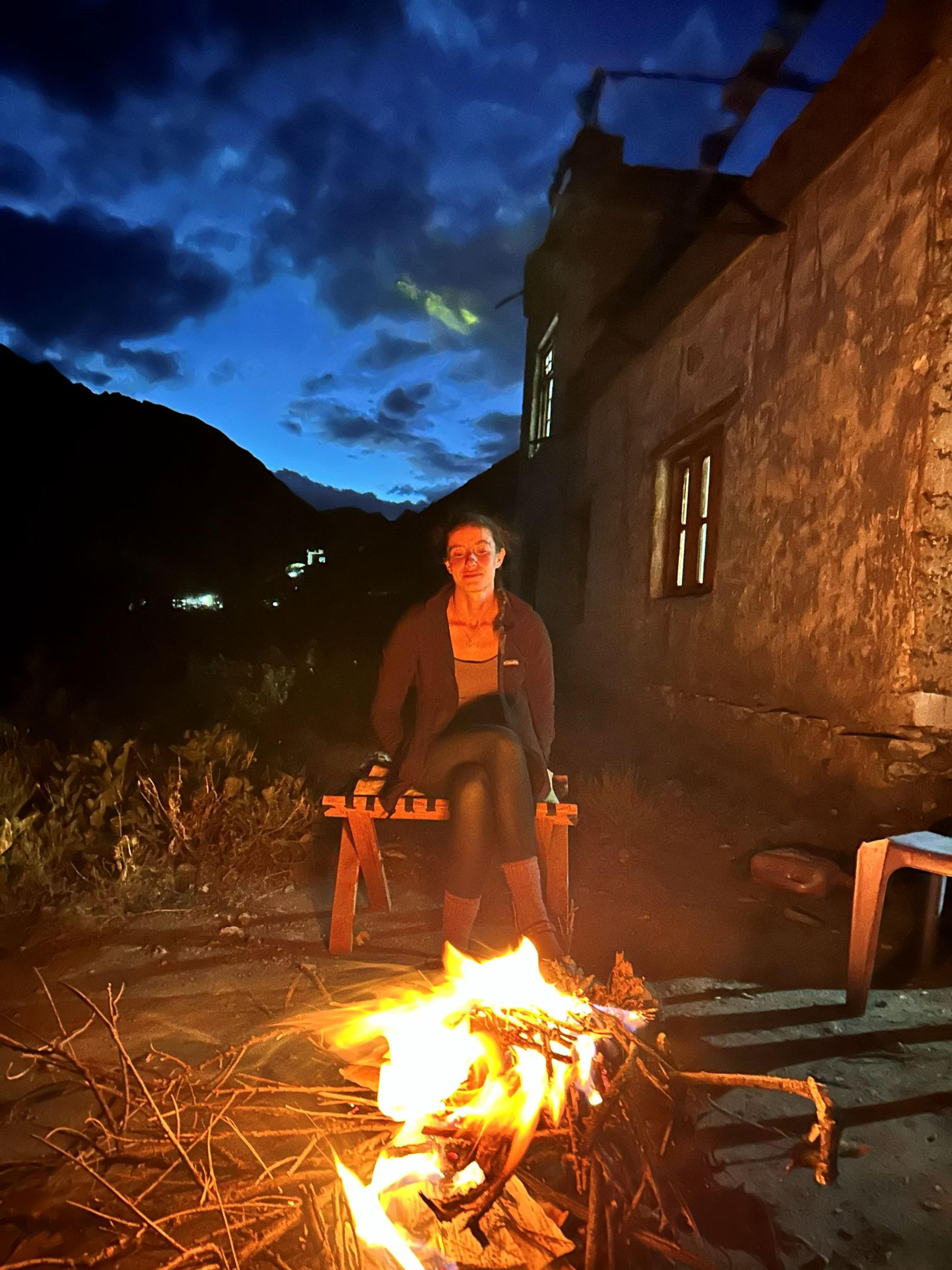 Trekker sitting by a small campfire outside a stone homestay at dusk, with dark mountains and a deep blue sky