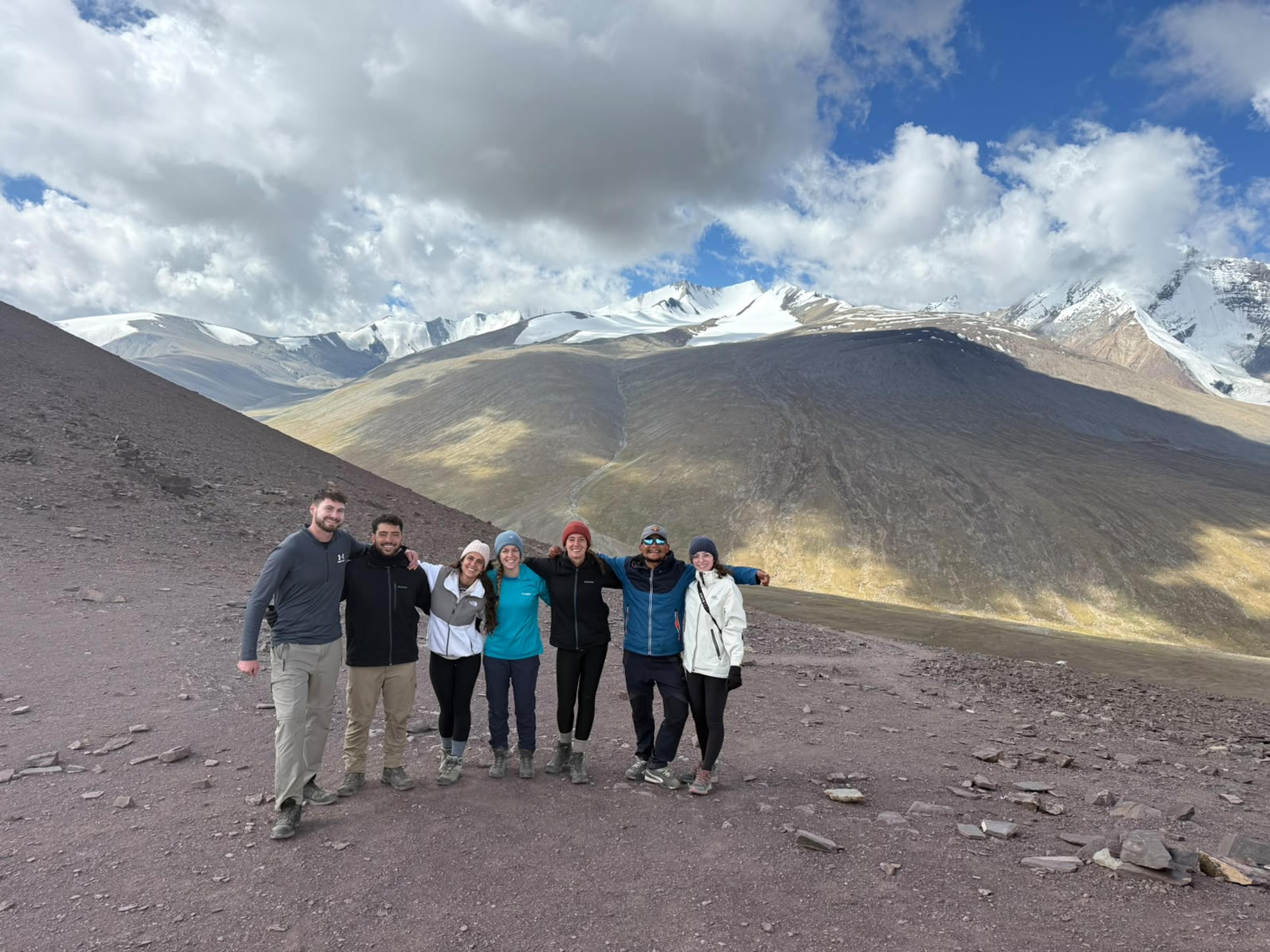Group of seven trekkers posing on a rocky slope above Nimaling, with snow-capped Kang Yatse peaks behind