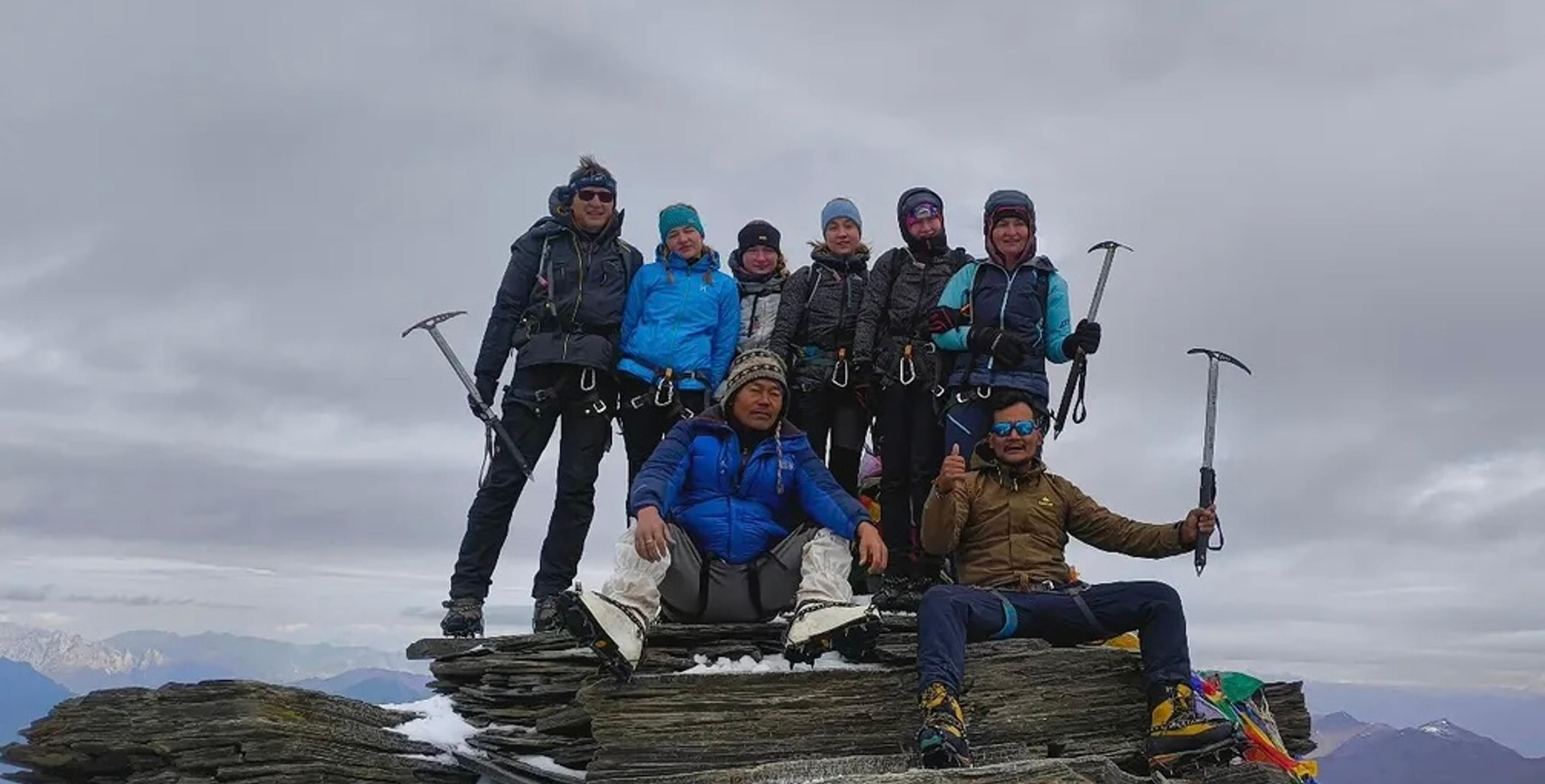A group of eight mountaineers posing with ice axes at a rocky Himalayan summit in Ladakh, with mountain ranges and overcast skies in the background