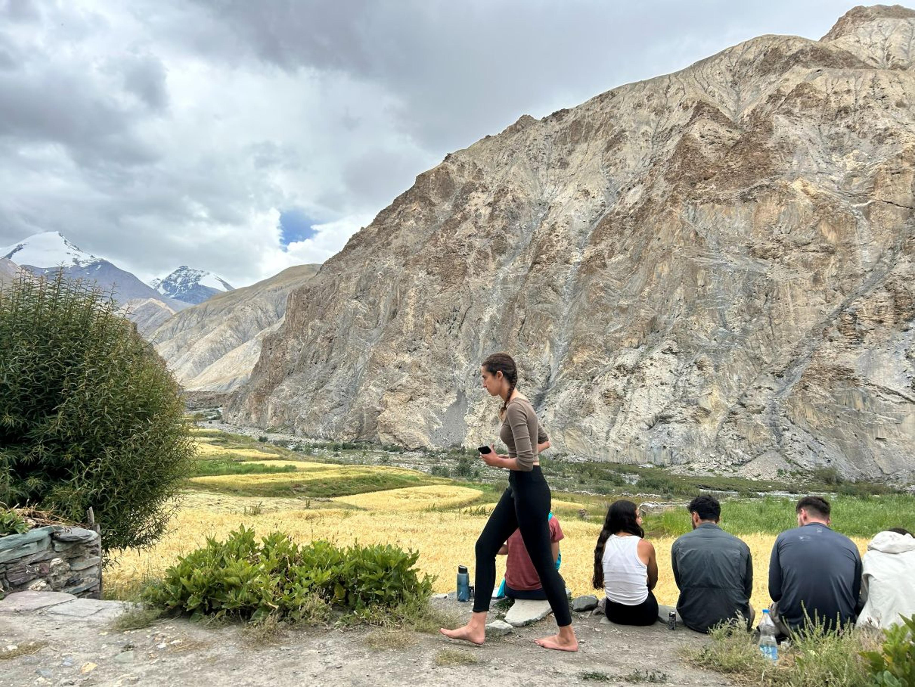 Trekkers resting beside golden barley fields in the Markha valley, with rocky cliffs and a snow-capped peak beyond