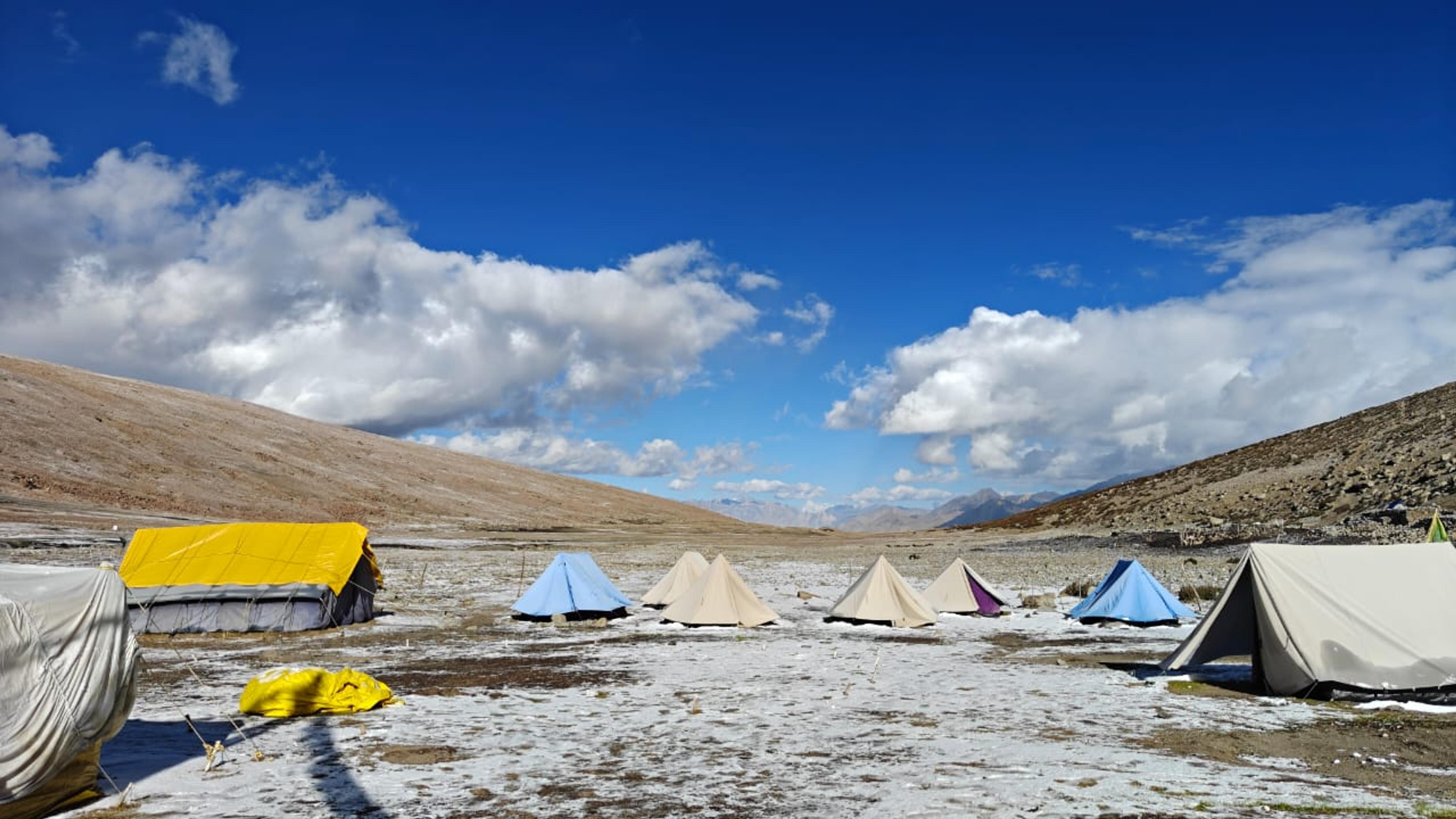 Colorful trekking tents set up on a snow-dusted open valley floor in Ladakh with barren hills and blue sky in the background