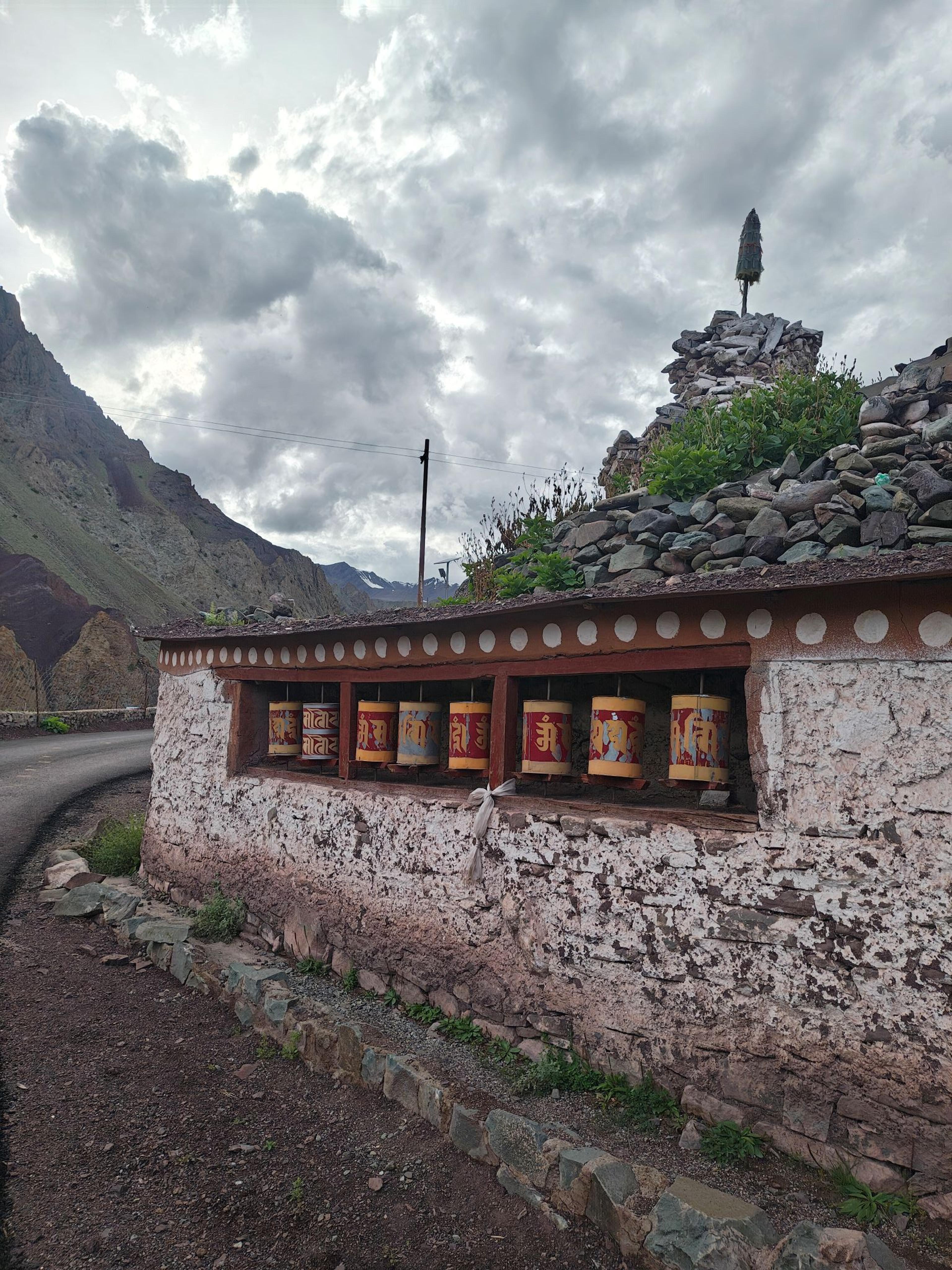 Row of red and yellow prayer wheels set into a whitewashed mani wall beside a road, under clouds with striped mountains