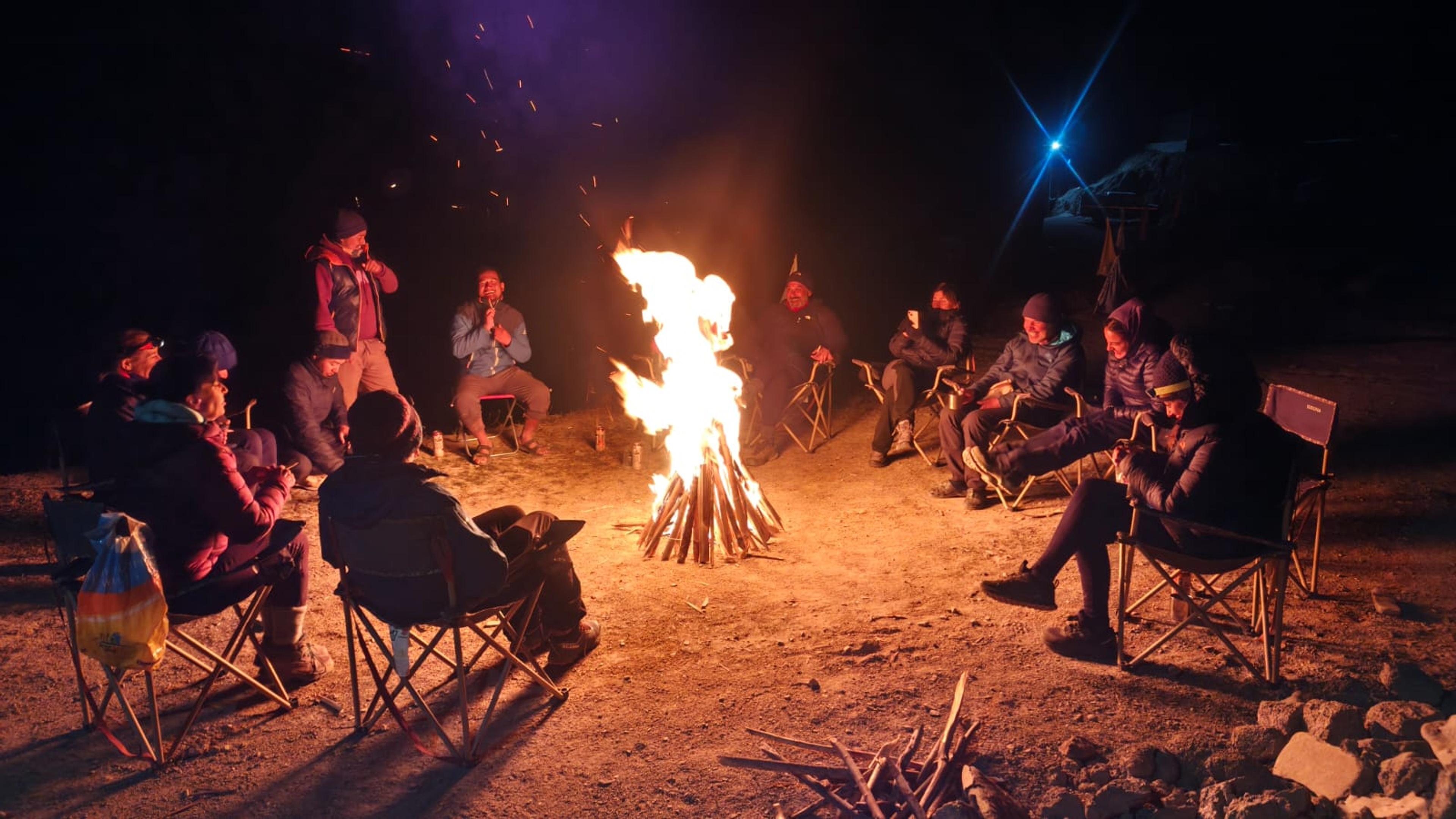 Group of travellers in down jackets sitting in camp chairs around a blazing bonfire at night, one singing into a mic