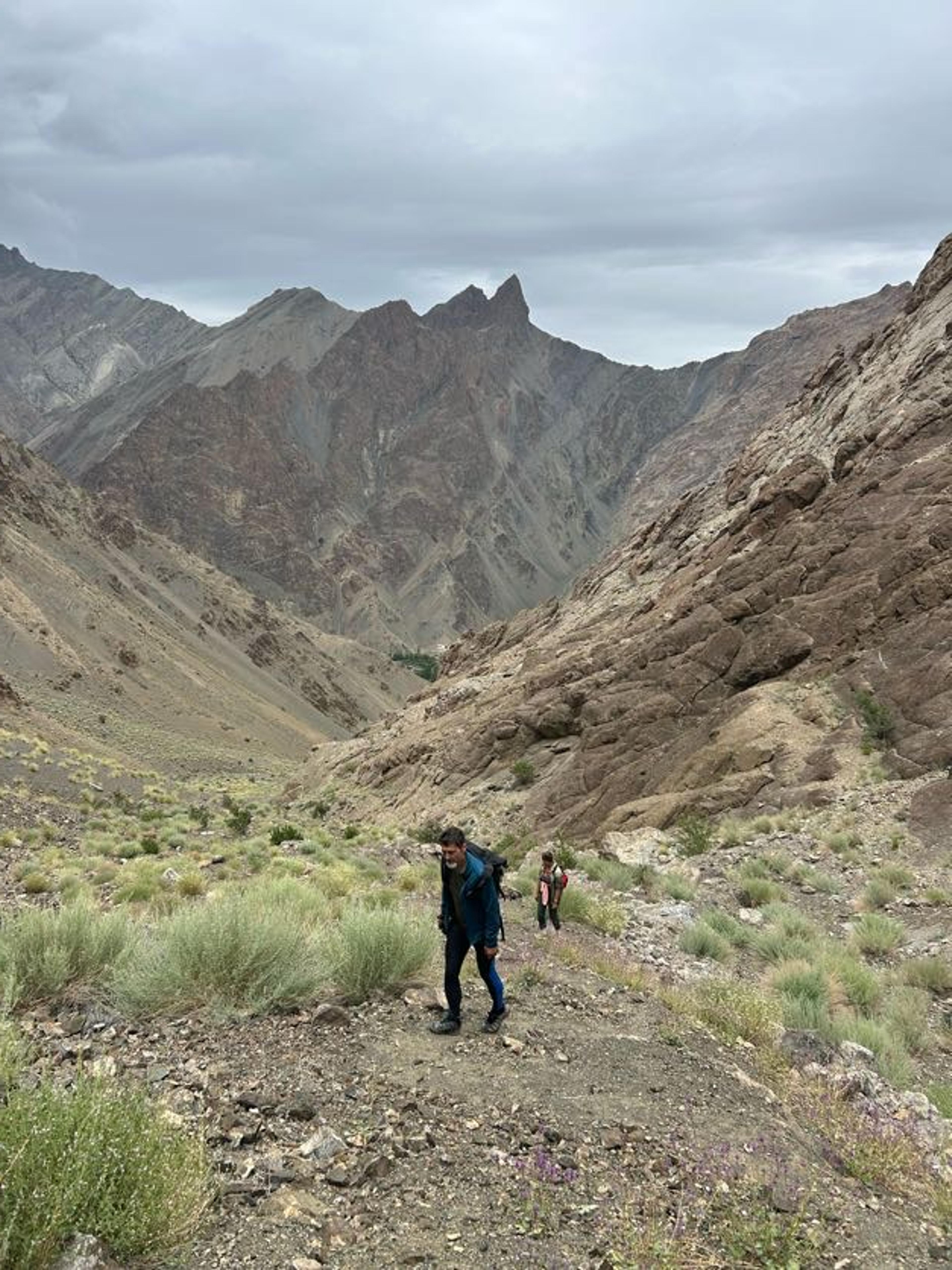 Two hikers climbing a steep, scrubby trail out of a valley, with jagged brown peaks under grey cloud behind