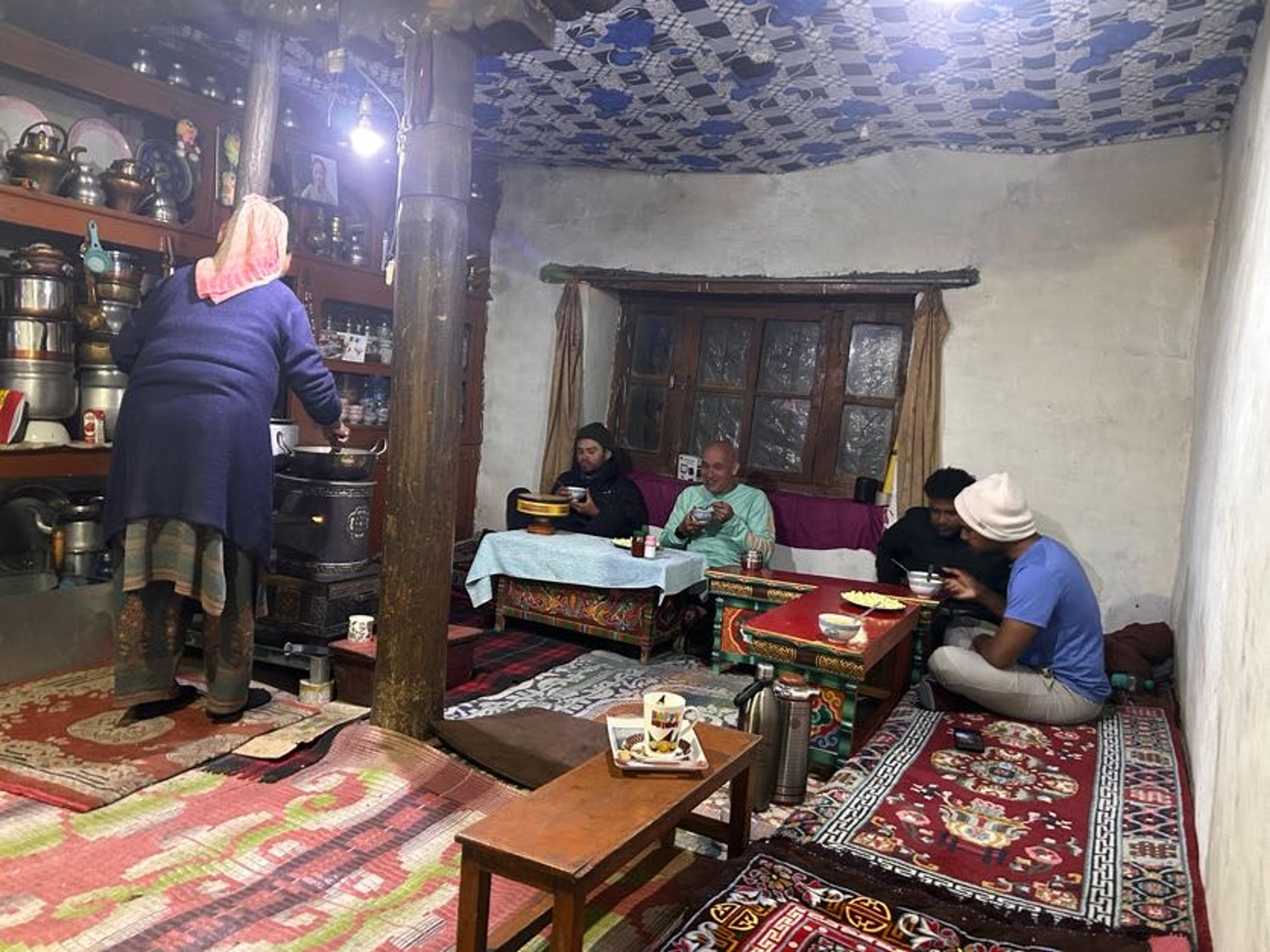 Travelers seated on floor rugs inside a traditional Ladakhi home being served tea by a local host, surrounded by traditional utensils, wooden furniture, and a patterned ceiling