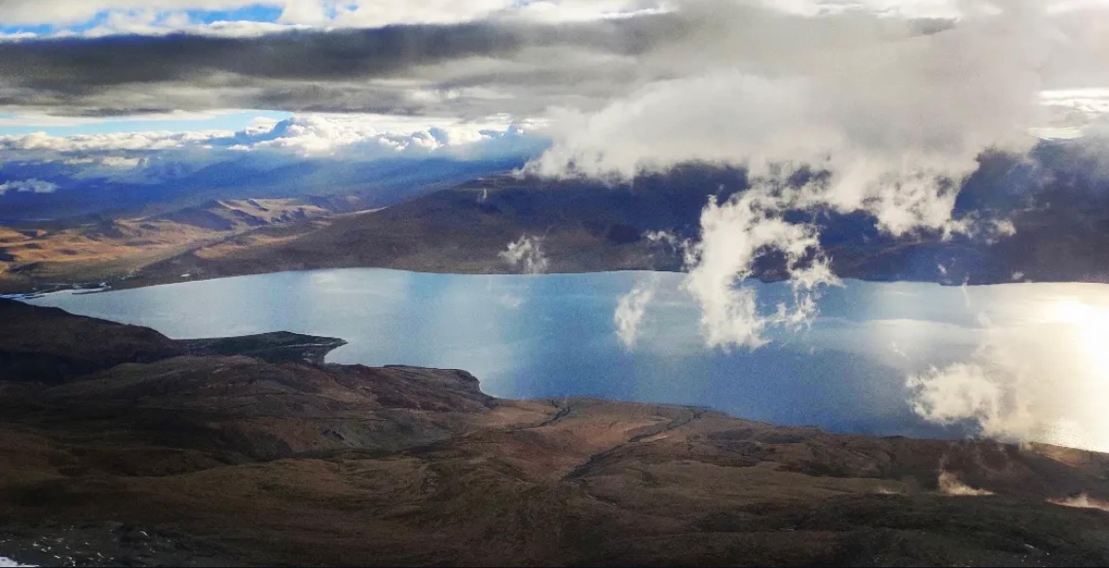 Bird's-eye view of Tso Moriri high-altitude lake surrounded by arid brown hills with dramatic clouds casting shadows on the blue water, seen from Mentok Kangri in Ladakh