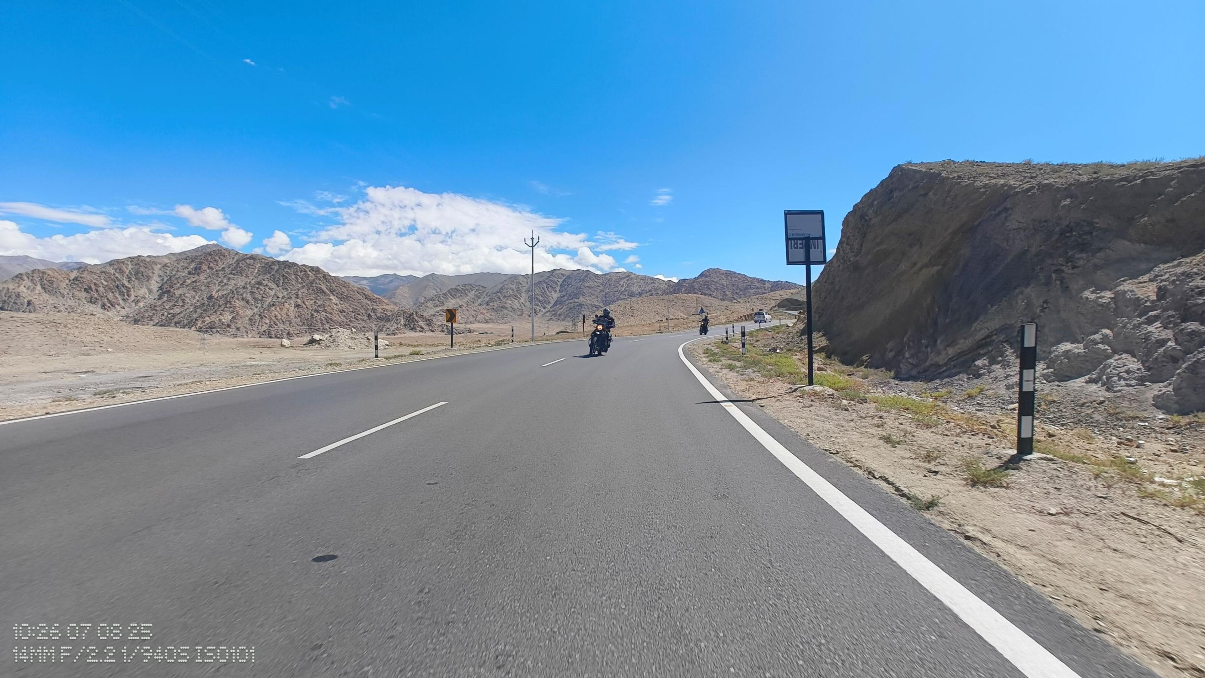 Two riders on Royal Enfield Himalayans approaching on a smooth, curving highway through arid brown mountains