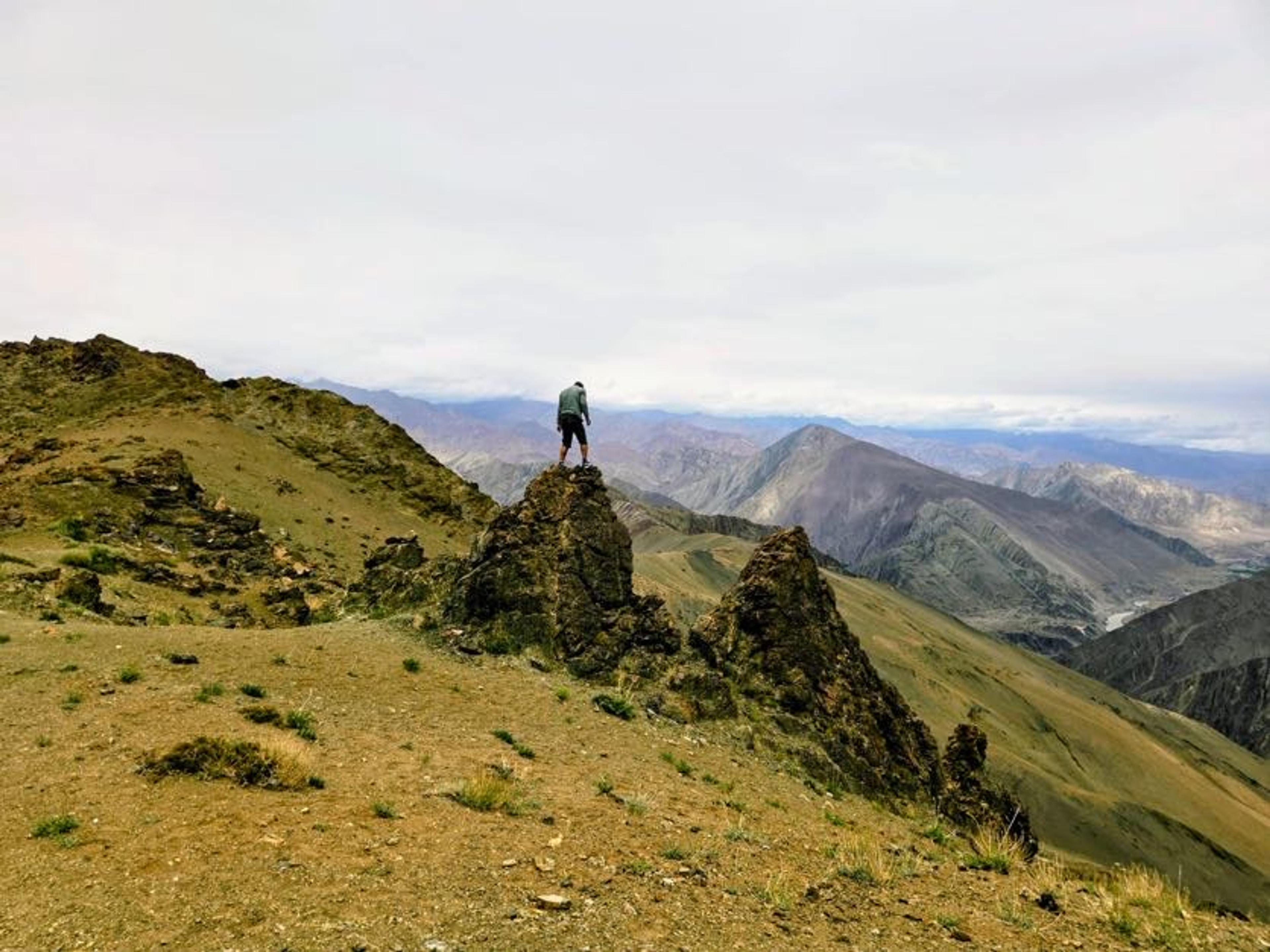 Hiker standing on a rocky outcrop on a high grassy ridge, looking over layered brown mountains and a river valley