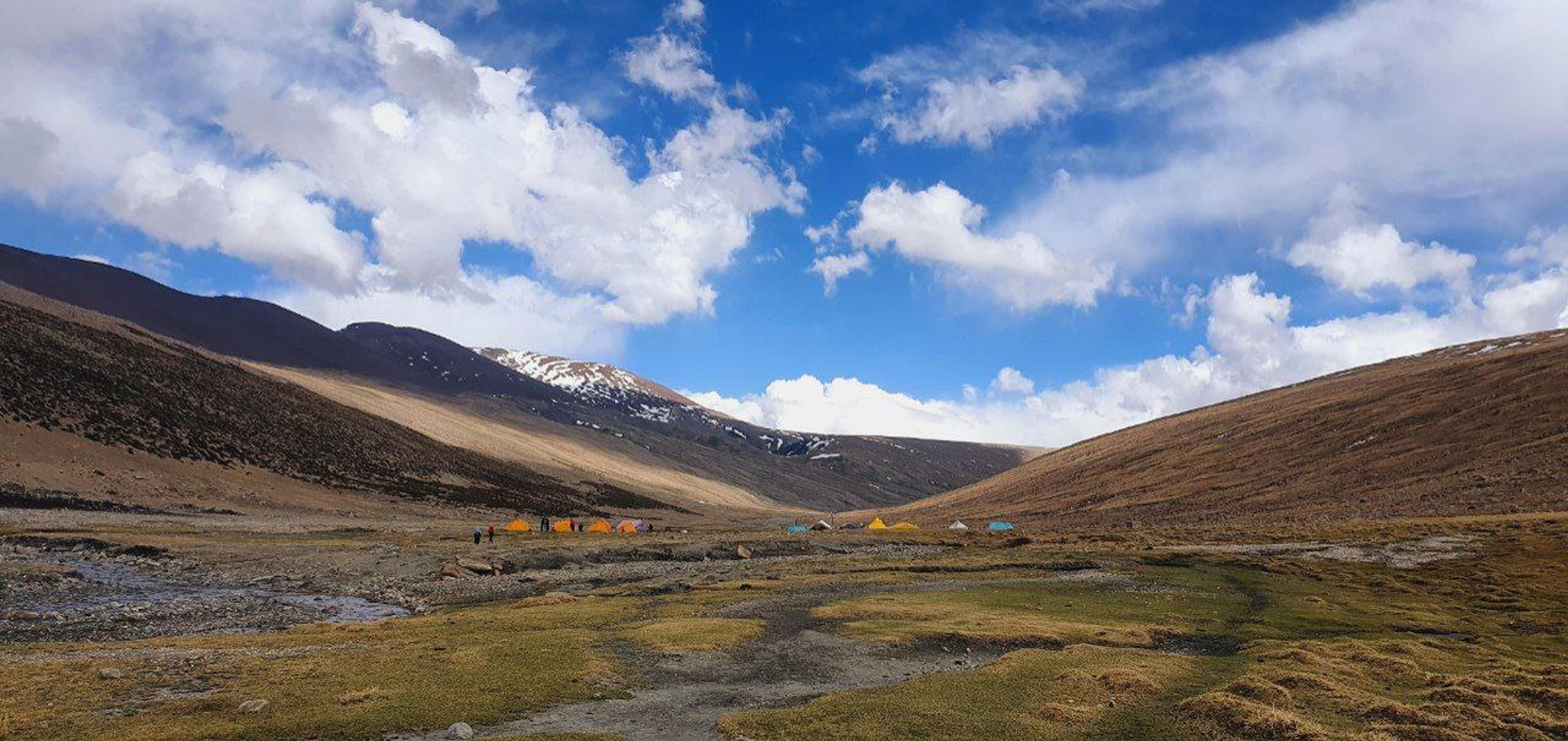 Colourful tents pitched on a high grassy meadow beside a stream, with snow-dusted ridges and blue sky