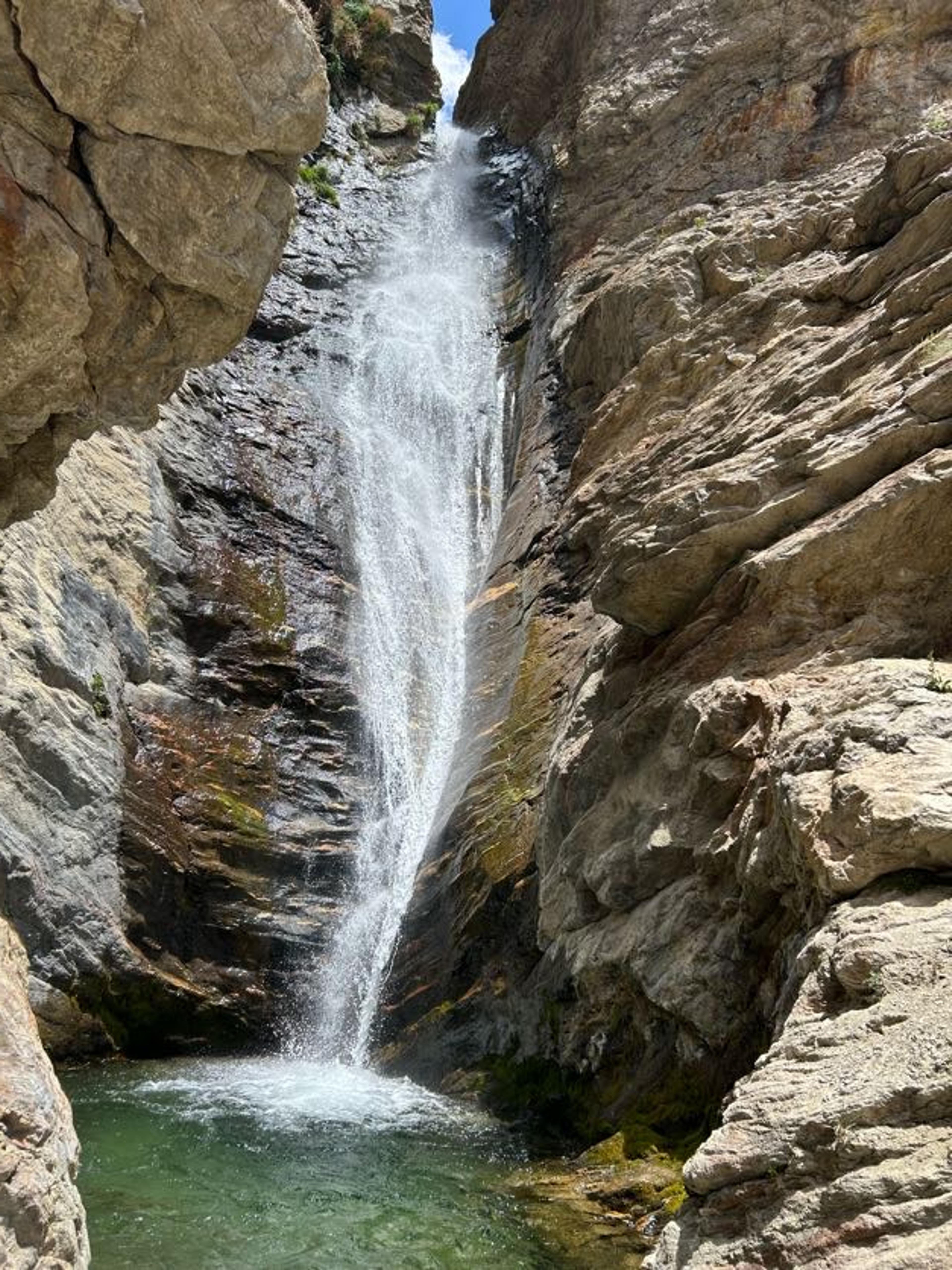 Waterfall plunging down a narrow rock gorge into a clear green pool, between grey and ochre cliffs