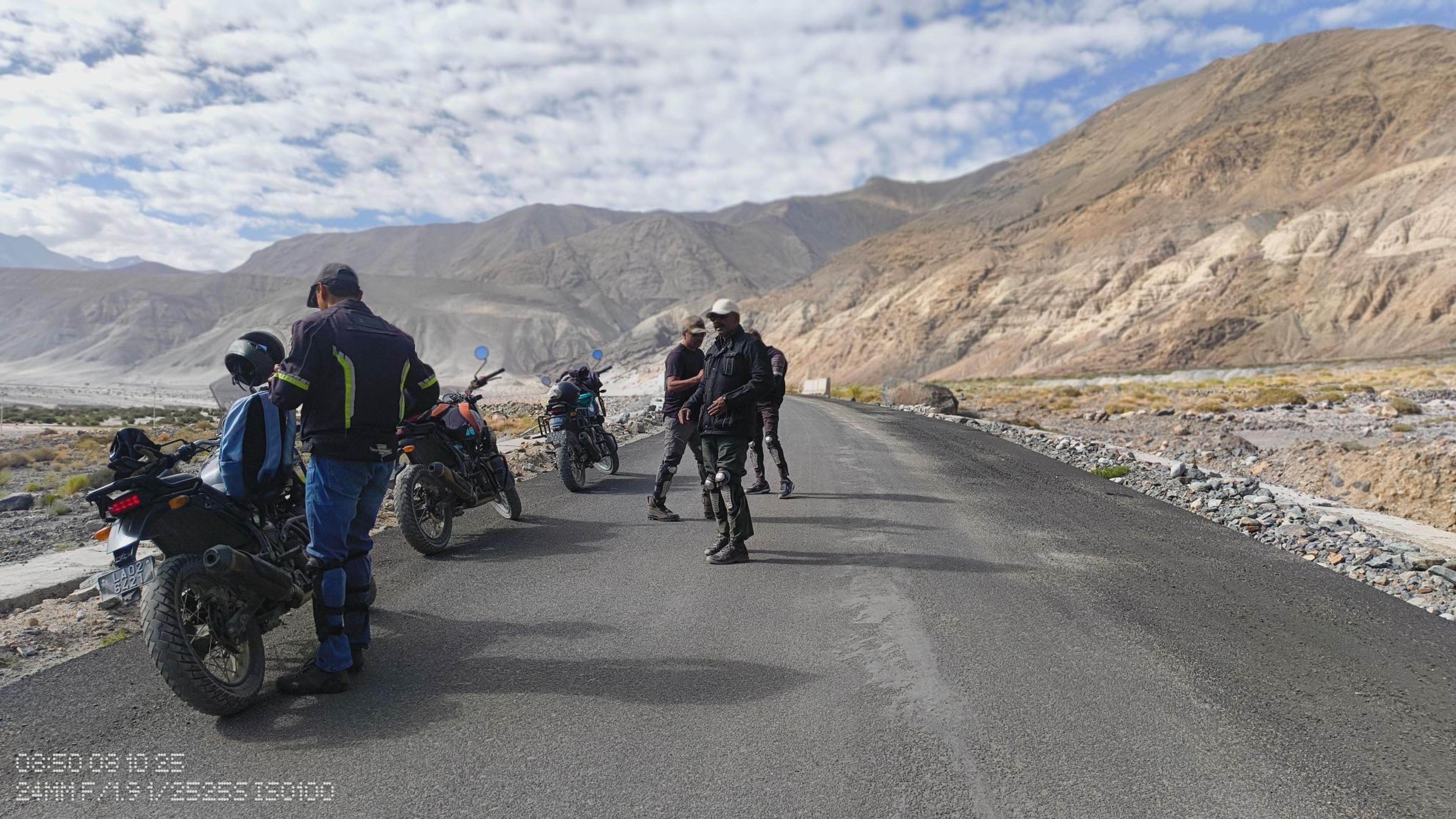 Riders stopped beside their Royal Enfield Himalayans on a mountain road in a wide, sandy river valley