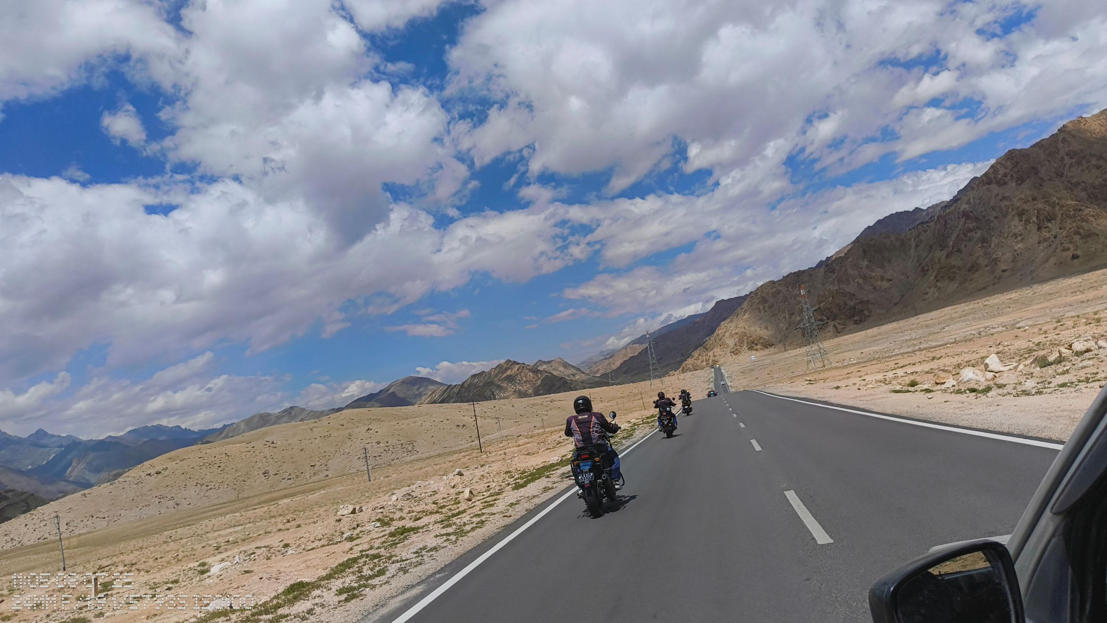 Riders on Royal Enfield Himalayans on a long, straight highway across an arid plain, under a cloudy blue sky