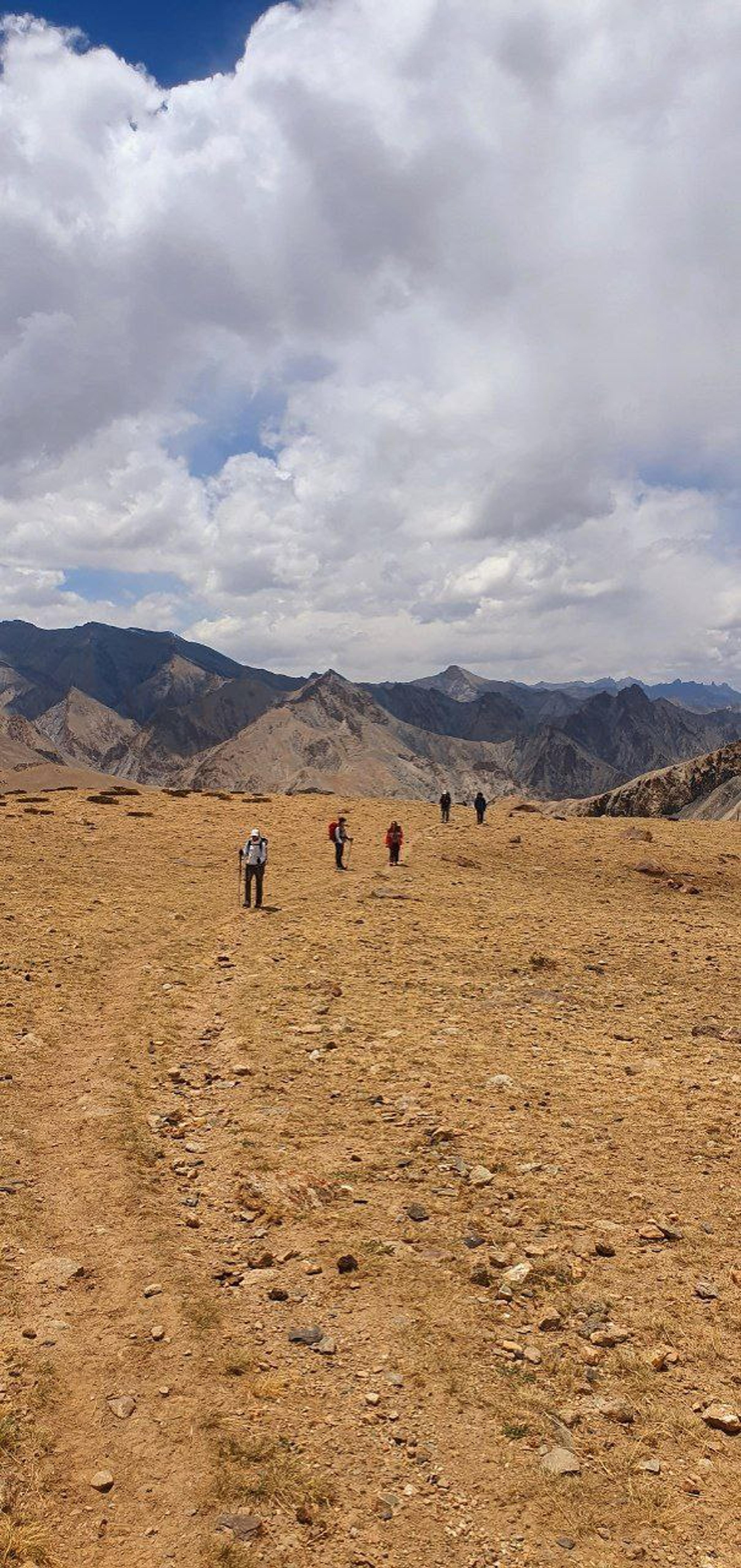 Trekkers hiking across a high-altitude plateau in Ladakh with dramatic mountain ranges and cloudy sky in the background