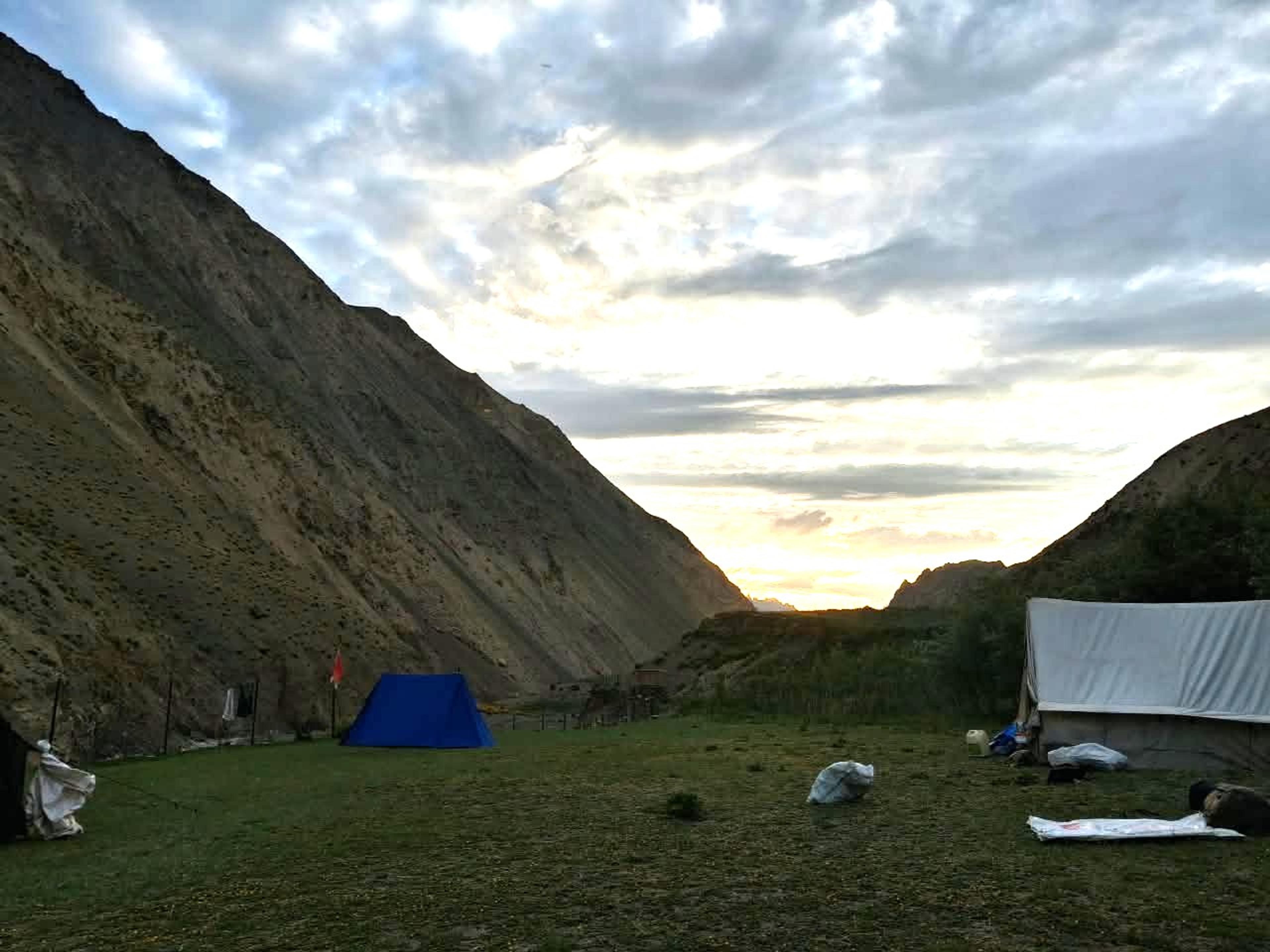 Tents on a grassy meadow at dusk in a deep valley between steep slopes, with the setting sun glowing through clouds