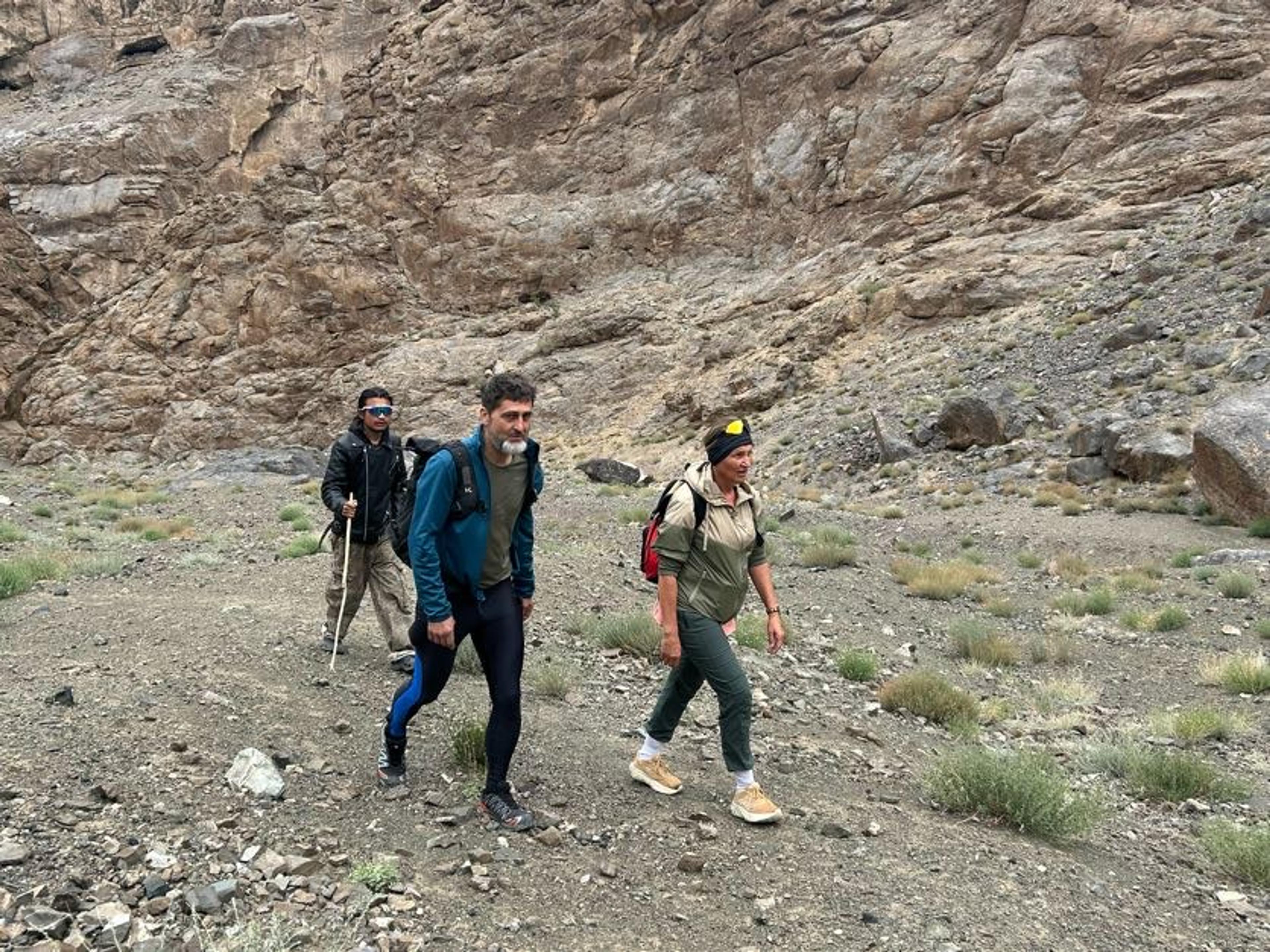 Three hikers with daypacks walking along a stony trail at the foot of steep brown rock walls