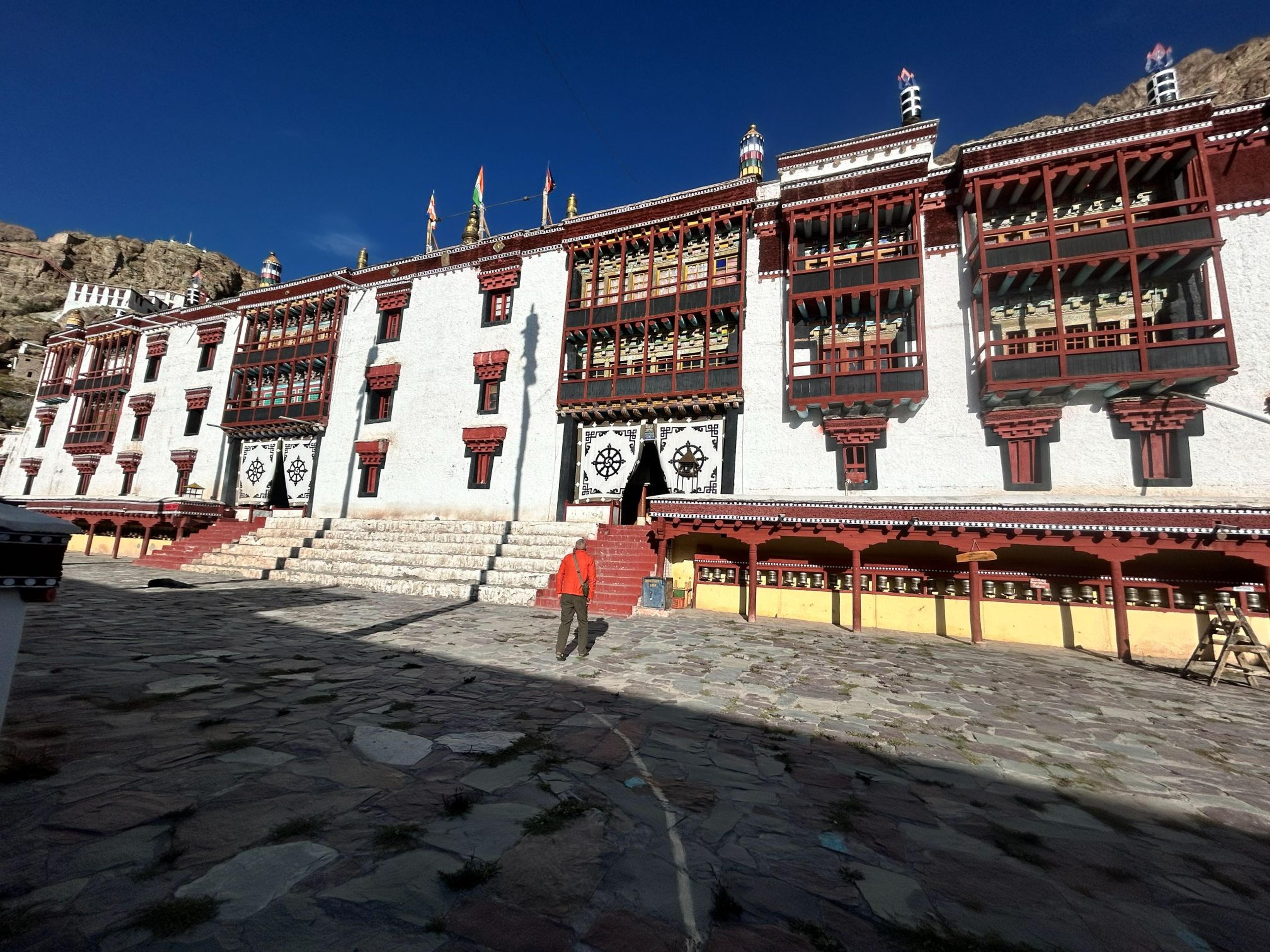 Visitor crossing the stone-paved courtyard of Hemis monastery, with white walls, red wooden balconies and prayer wheels