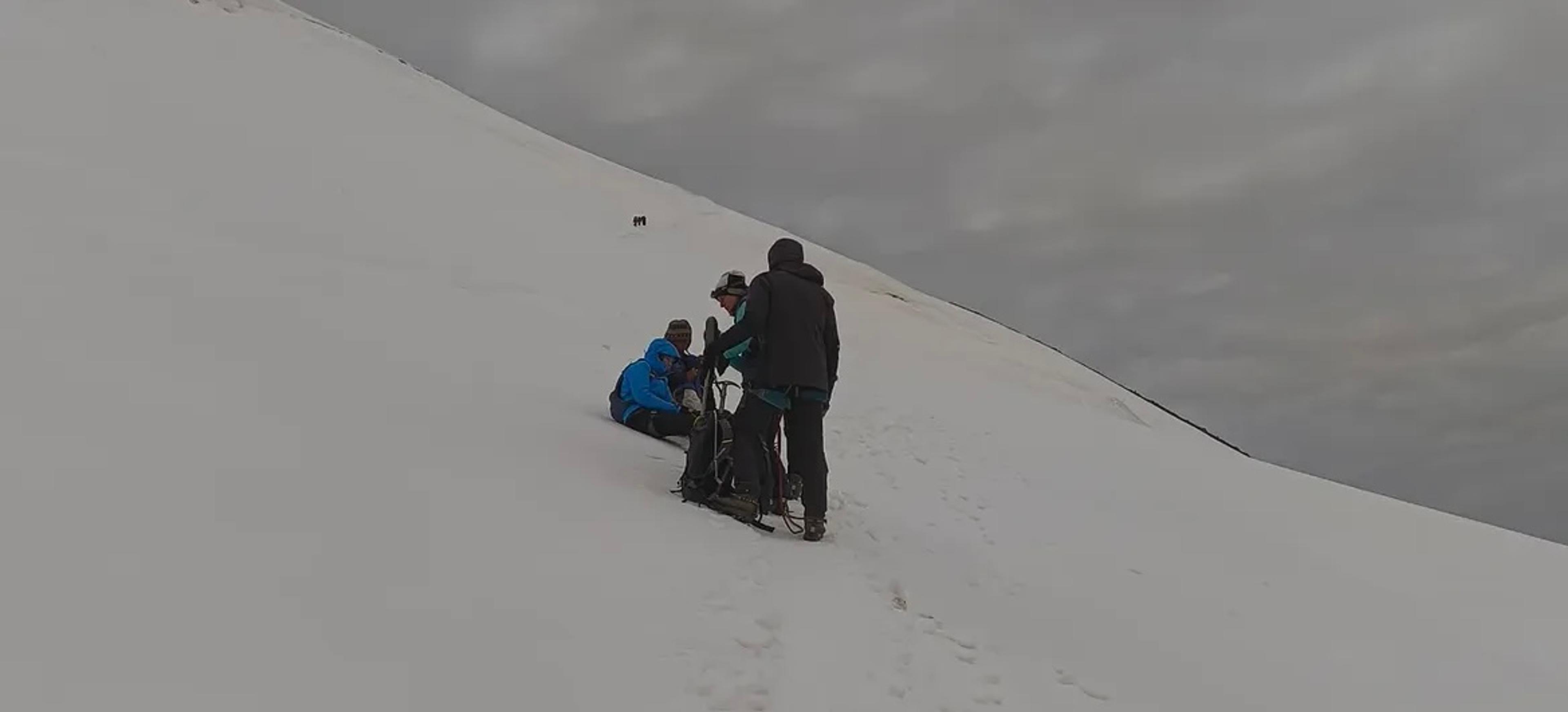 Three mountaineers in winter gear resting and assisting each other on a steep snow slope, with footprints in the snow and an overcast sky, Himalayan peak in Ladakh