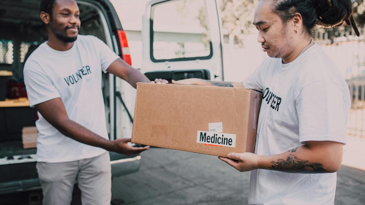 Two volunteers distribute a box of medicine from a van, emphasizing charity and teamwork.
