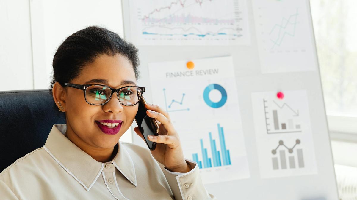 Confident businesswoman wearing glasses studies financial charts while talking on smartphone.