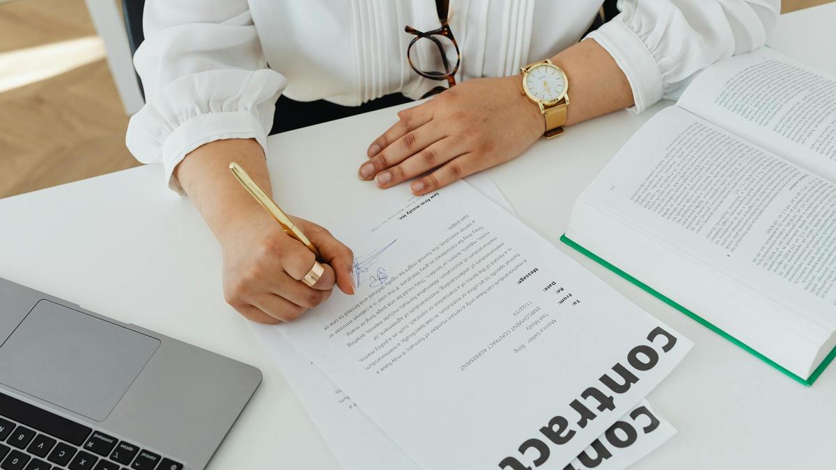 Close-up of a businesswoman signing a contract at her desk, with a laptop and book.